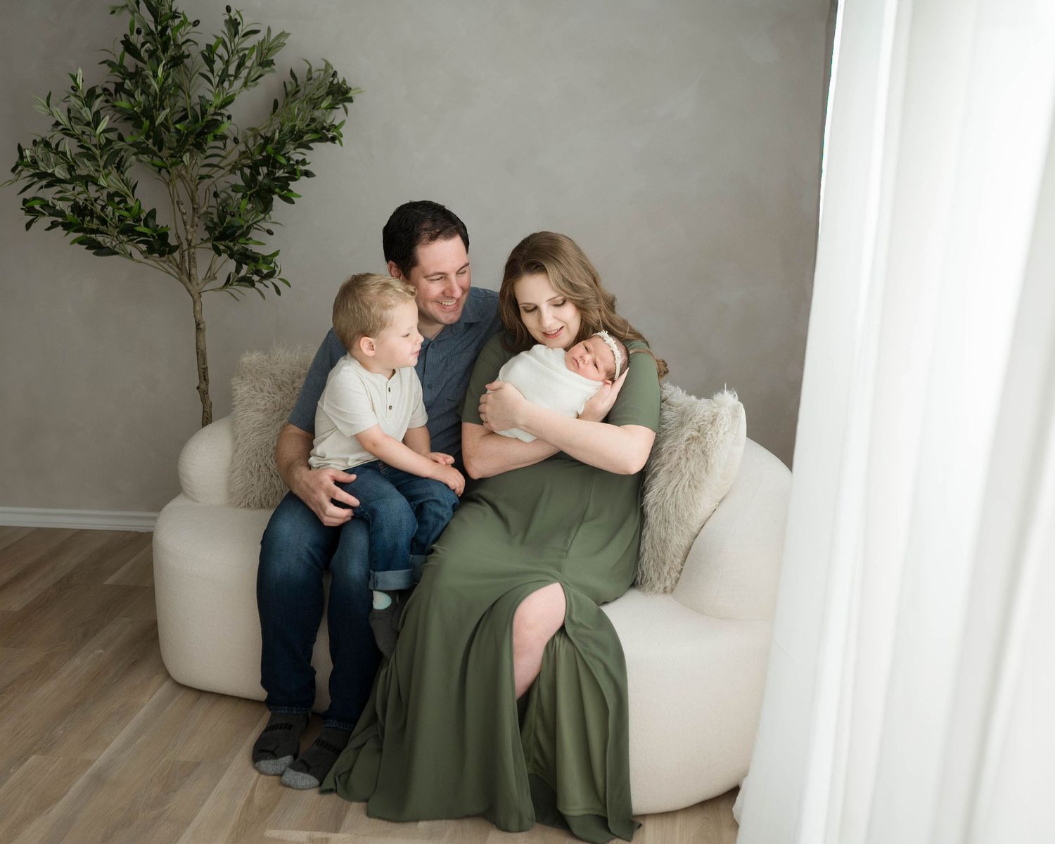 Family of four sitting on a cream sofa, parents embracing newborn baby with toddler son beside them.