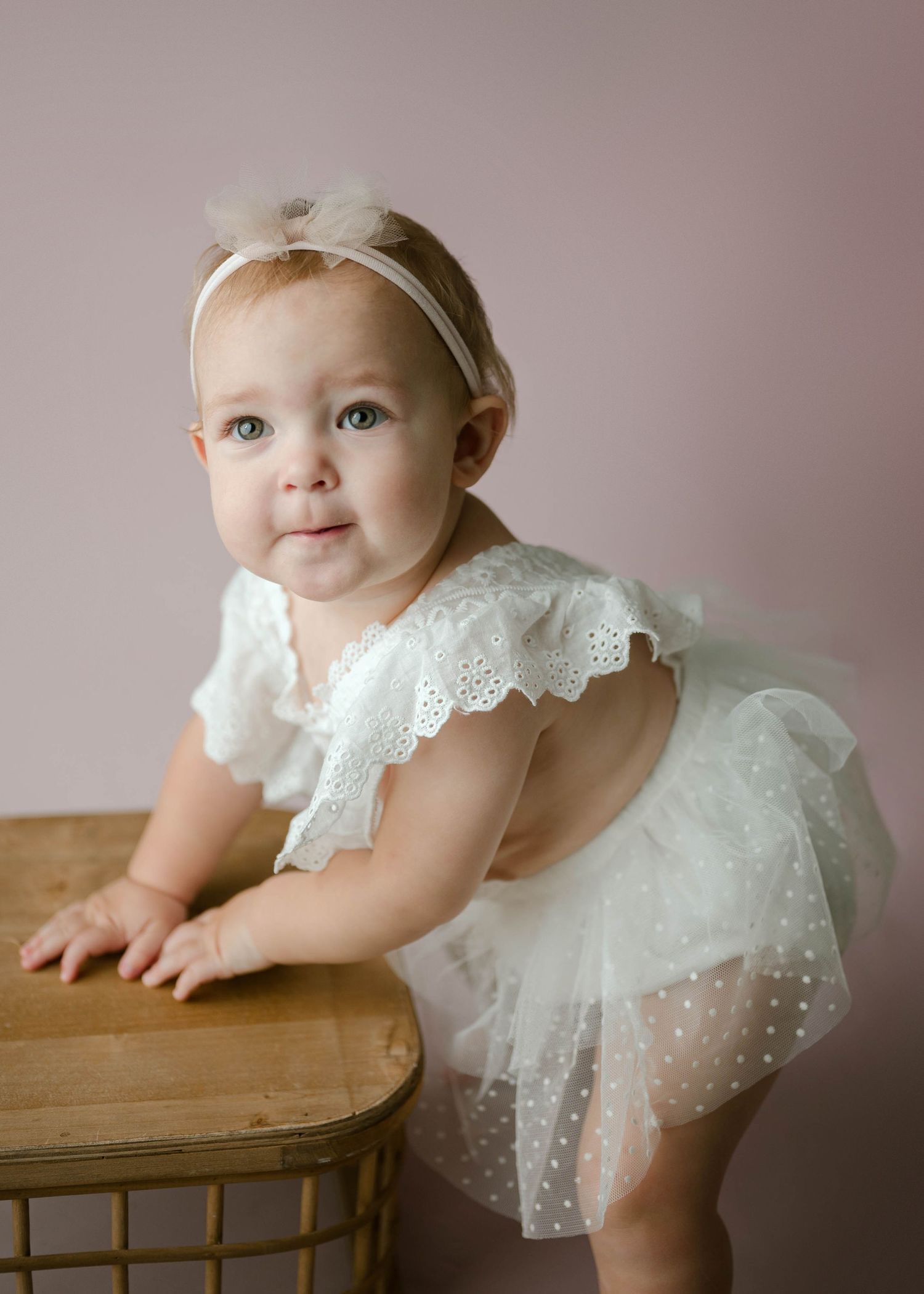 Baby girl in white lace tutu dress and bow headband leaning on wooden stool during studio portrait session.
