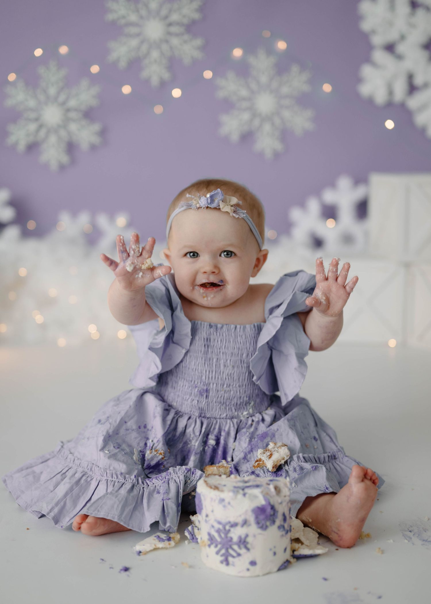 Laughing baby girl in purple dress holds up frosting-covered hands beside smashed snowflake birthday cake.