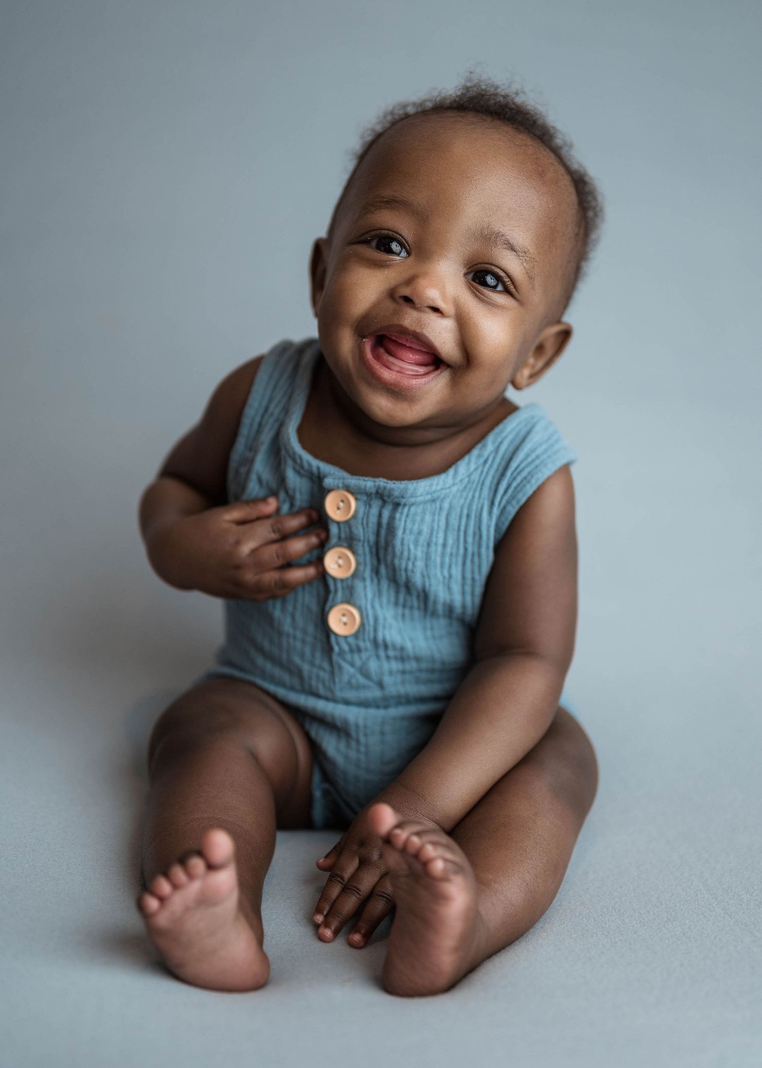 Joyful baby in teal romper laughing while sitting on gray background.