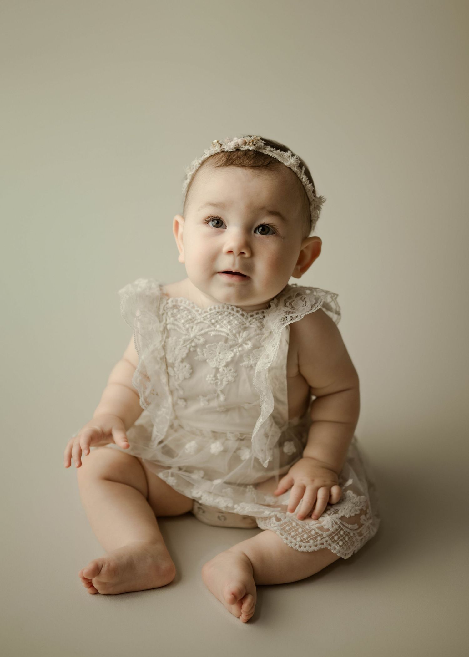 Sweet baby girl in ivory lace romper and headband sitting on studio floor against sage background.