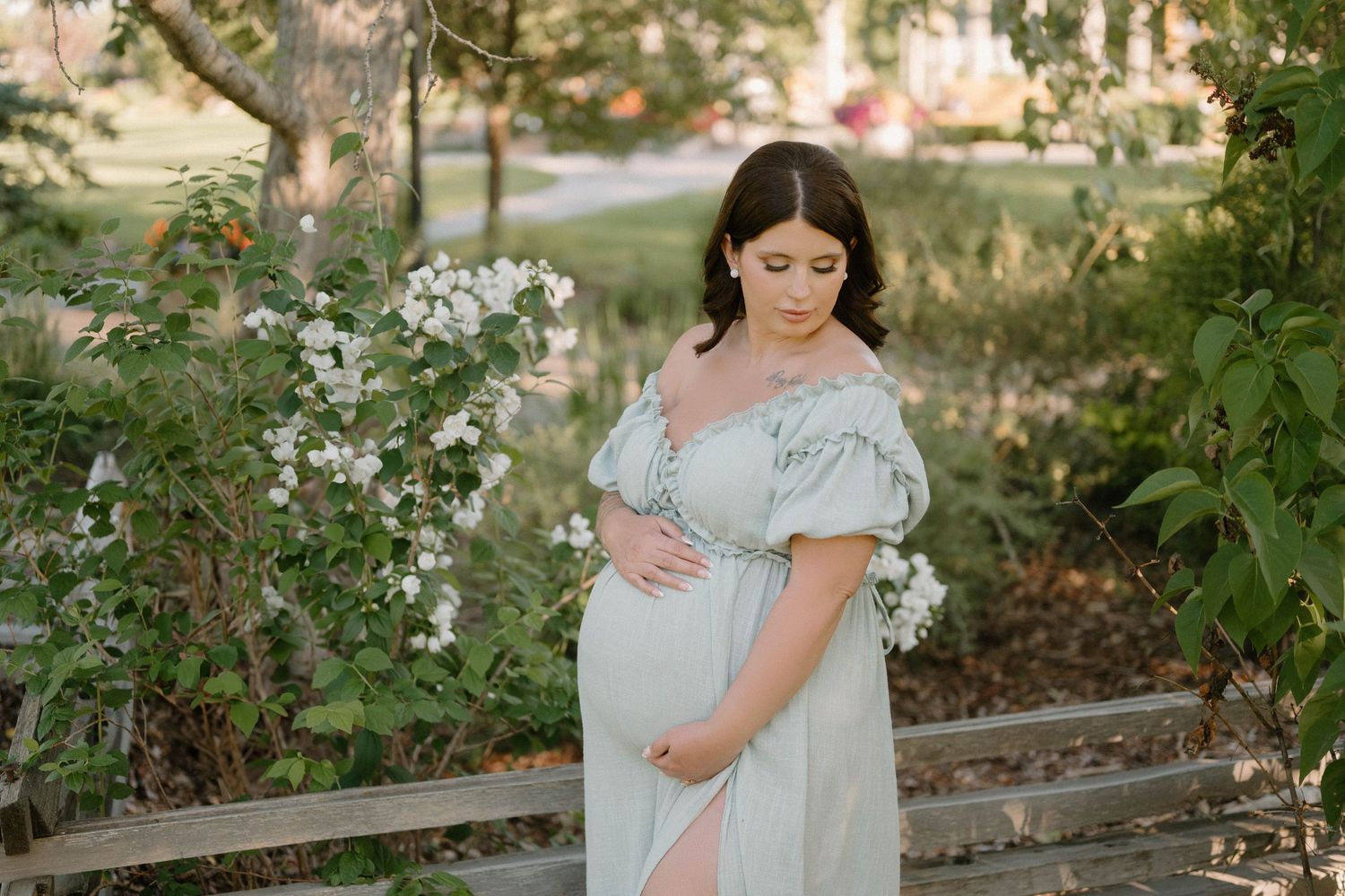 Pregnant woman in mint off-shoulder gown cradling belly outdoors near white flowering bushes.