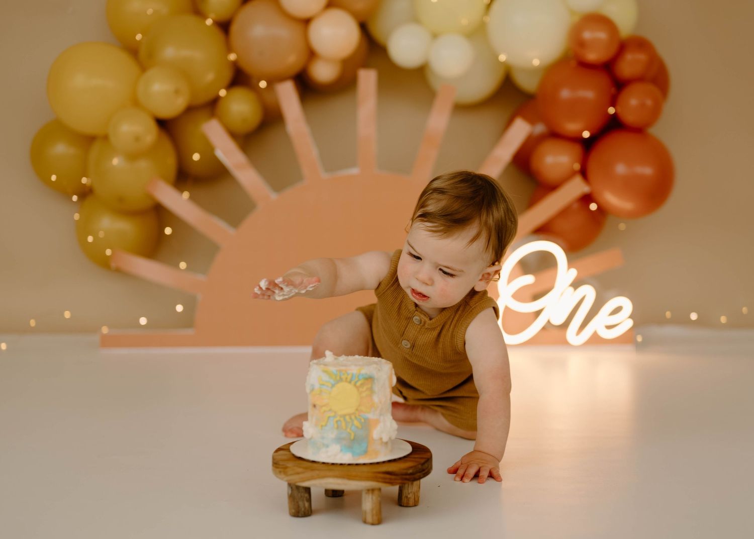 Baby boy in brown romper reaching for birthday cake with colorful balloon sun backdrop.