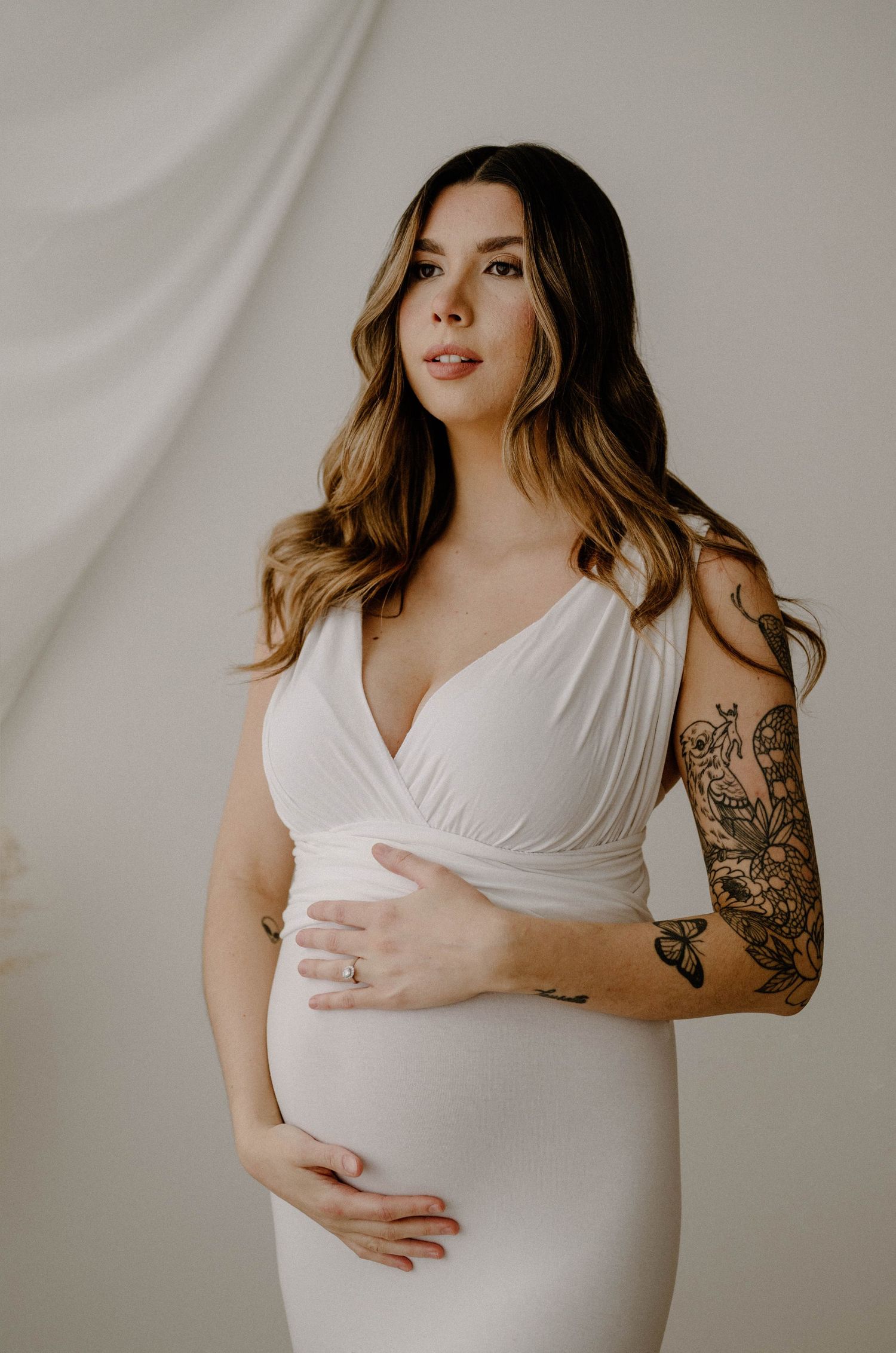 Close-up of tattooed pregnant woman in white wrap dress cradling her baby bump.