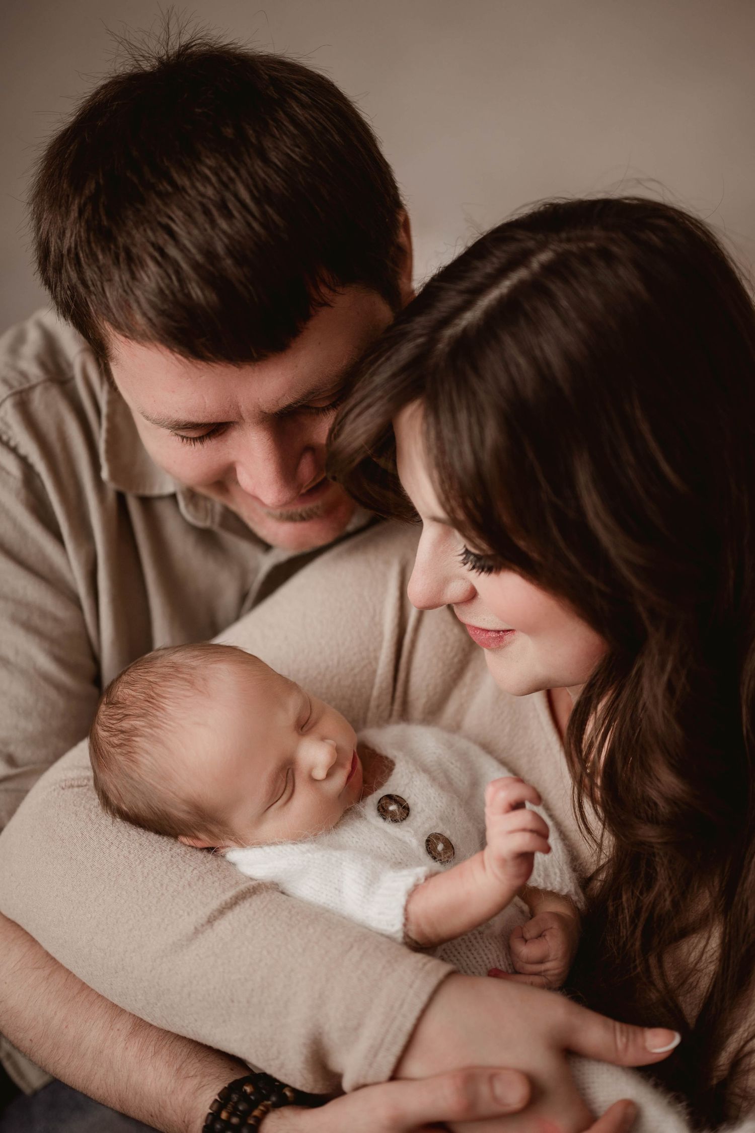 Young couple smiling and gazing lovingly at their sleeping newborn baby wrapped in a white blanket.