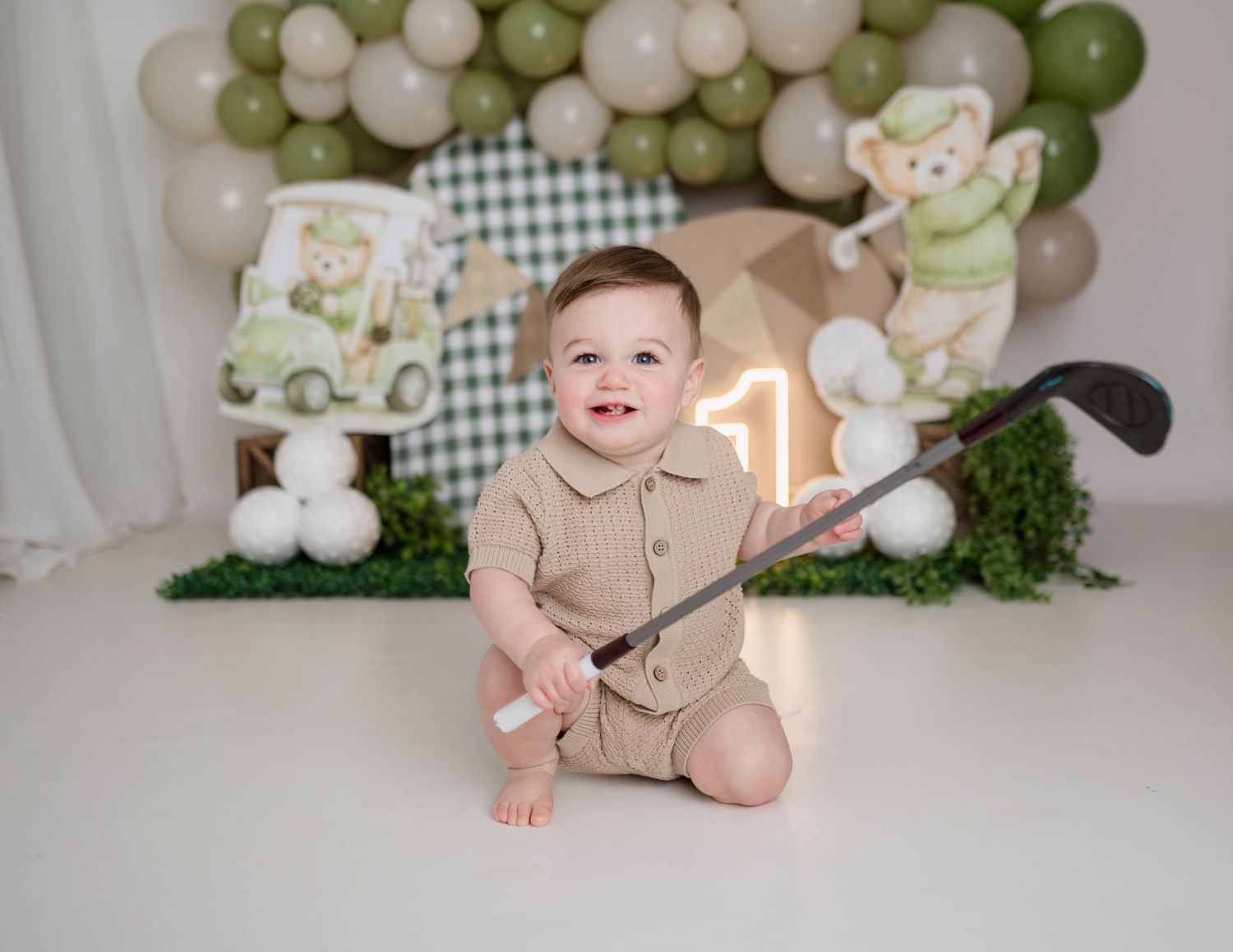 Smiling baby boy in beige romper holds golf club at a golf-themed first birthday cake smash photo session.