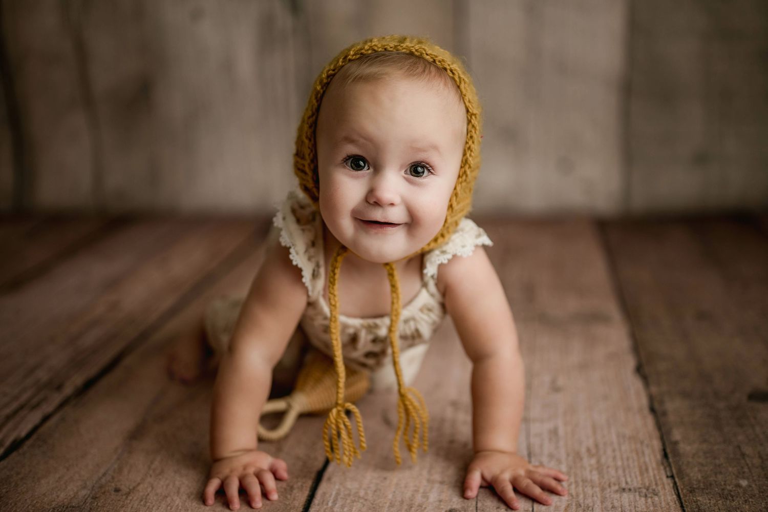 Smiling baby in cream romper and mustard knit bonnet crawls on rustic wooden floor in studio portrait.