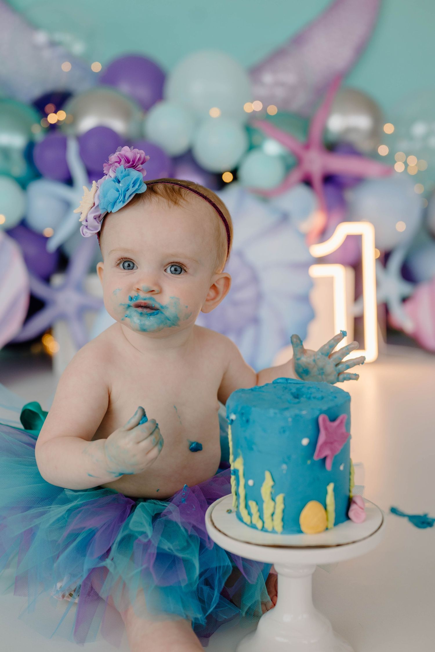 One-year-old baby girl with blue frosting on face sits beside mermaid-themed first birthday cake smash setup.