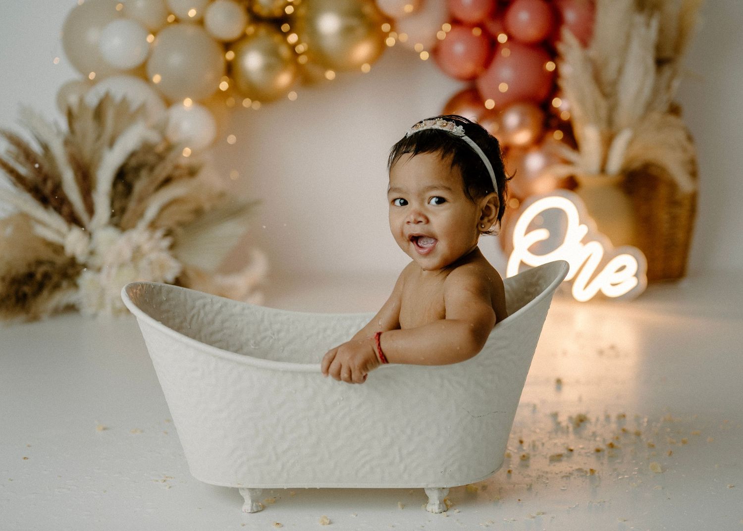 Smiling baby girl sits in white clawfoot tub at boho first birthday bath session with balloon backdrop.