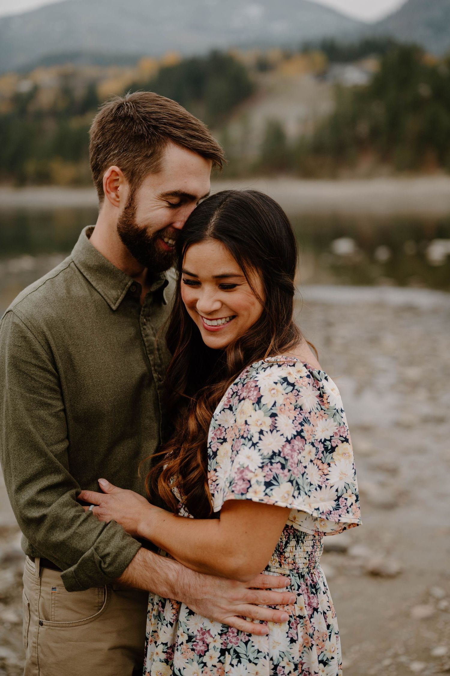 Couple embraces and smiles together outdoors near a mountain lake during a fall engagement session.