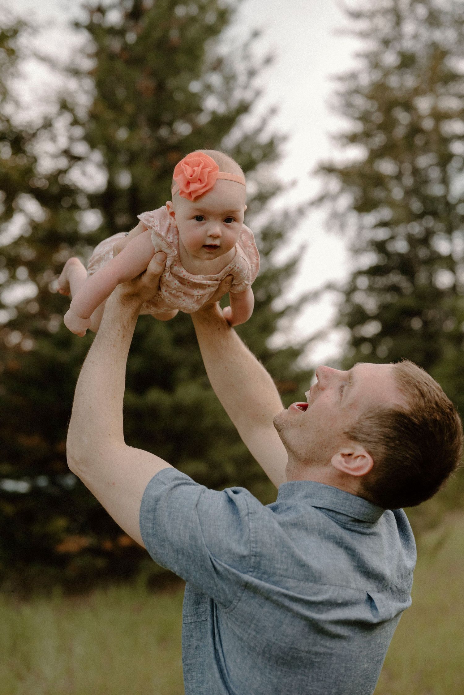 A father joyfully lifts his baby girl above his head in an outdoor setting.