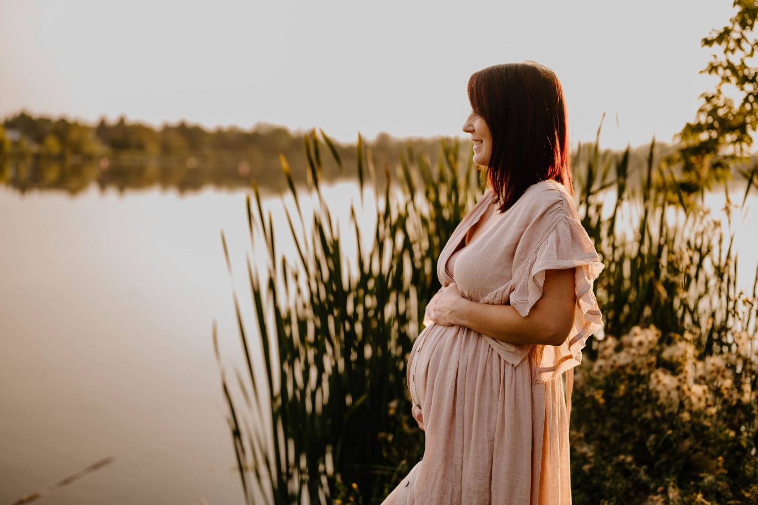 Pregnant woman in blush pink dress standing peacefully by Telford lake at golden sunset.
