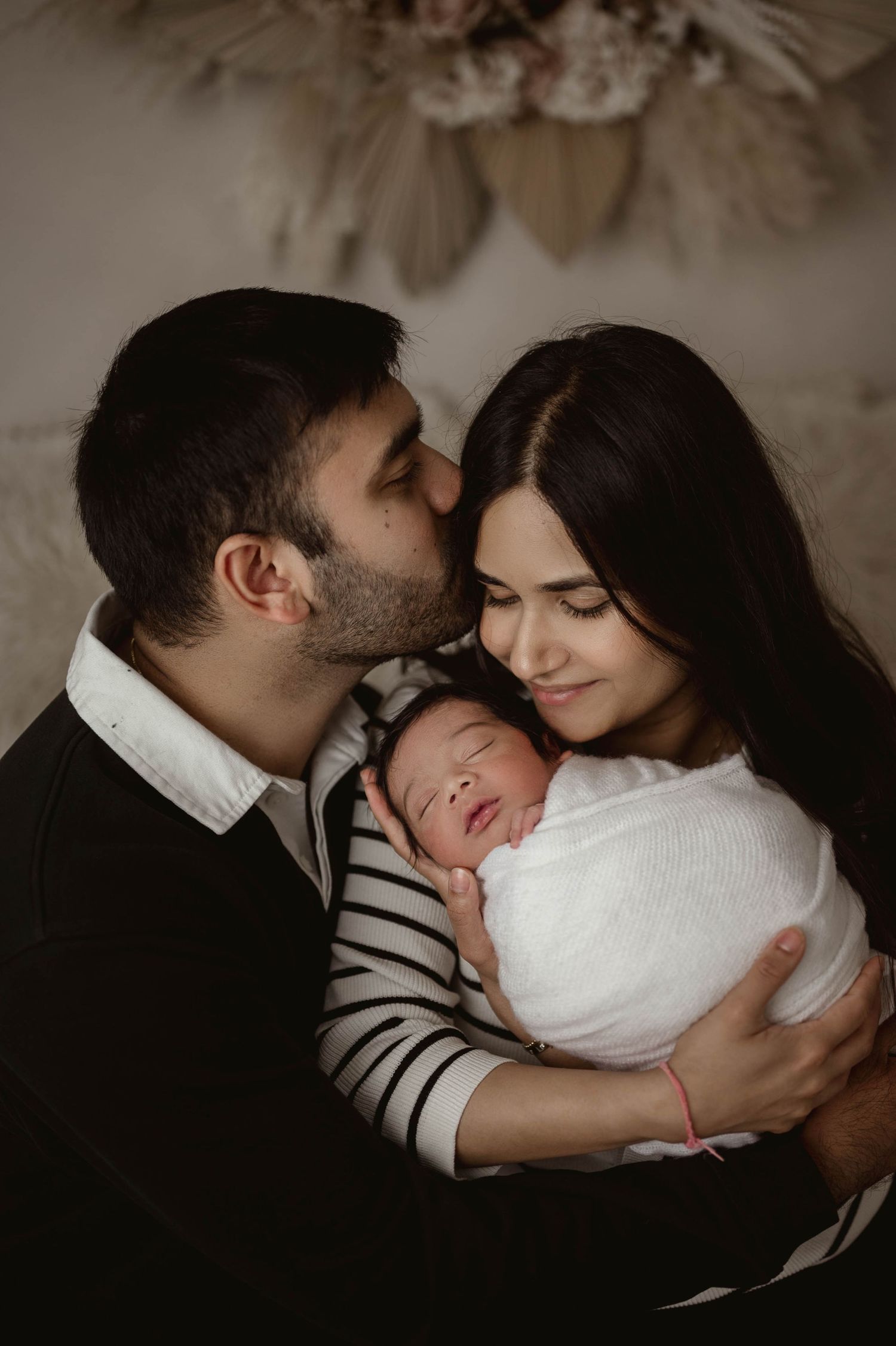 A couple tenderly holds their swaddled newborn baby, with the man kissing the woman's temple in a warm studio portrait.