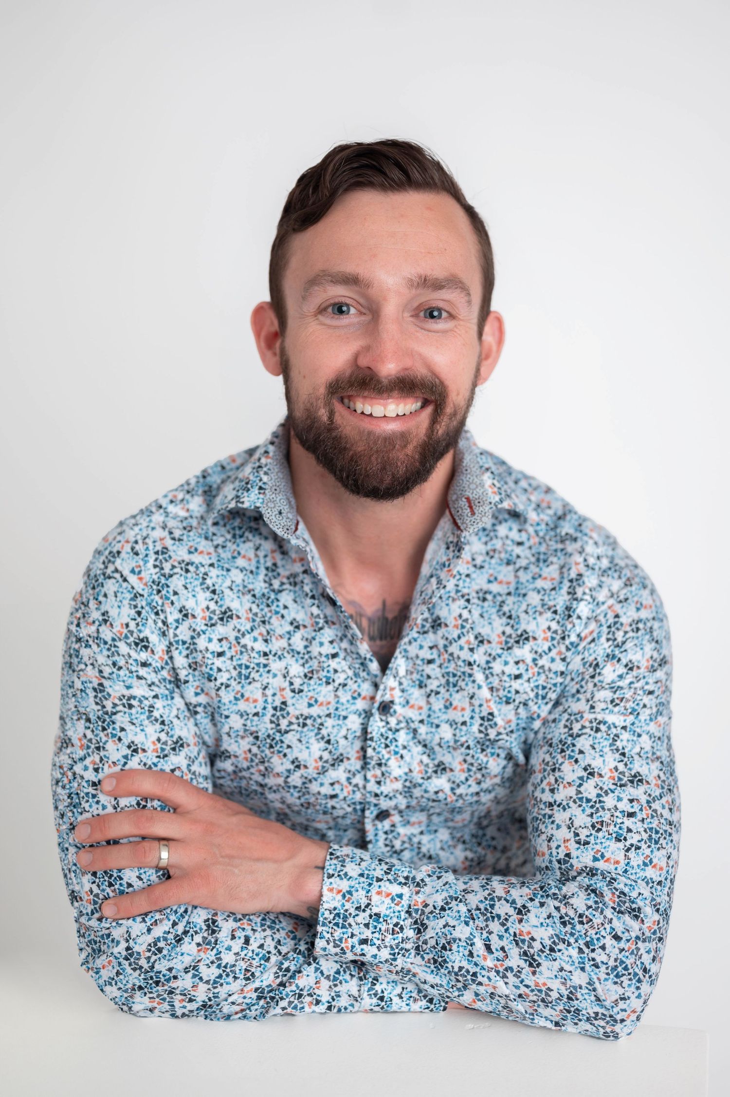 Smiling bearded man with crossed arms wearing a blue floral shirt against a white background.