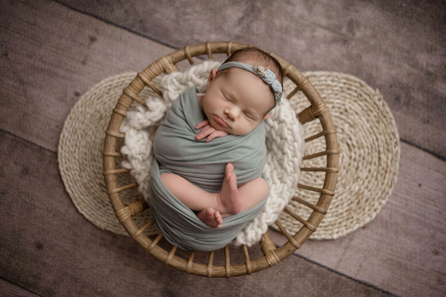 Newborn baby swaddled in sage green wrap sleeping peacefully in a wicker basket with white flokati fur.