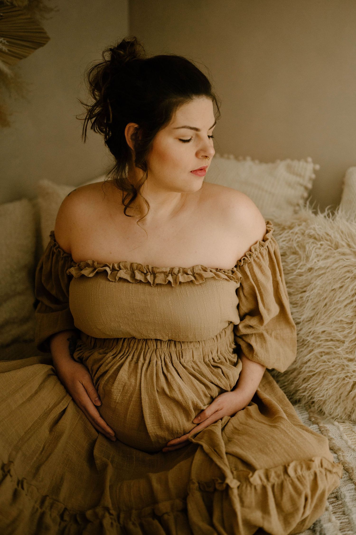 Pregnant woman in tan off-shoulder ruffle dress sitting on bed cradling bump.