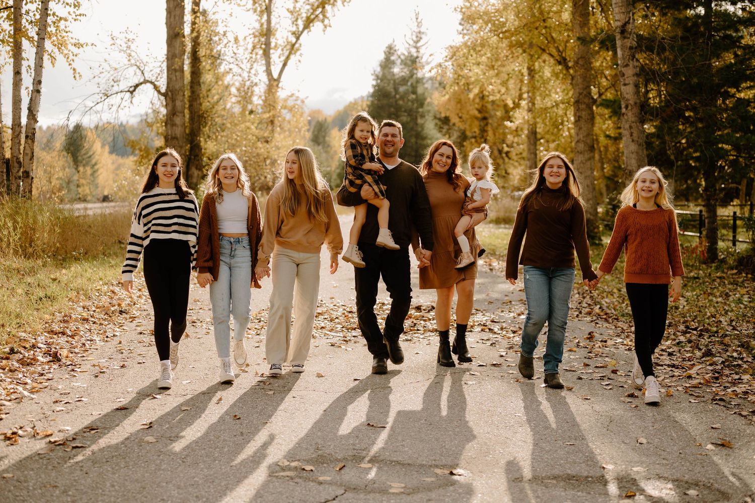 Family of nine walking together on a sunlit path surrounded by fall foliage.