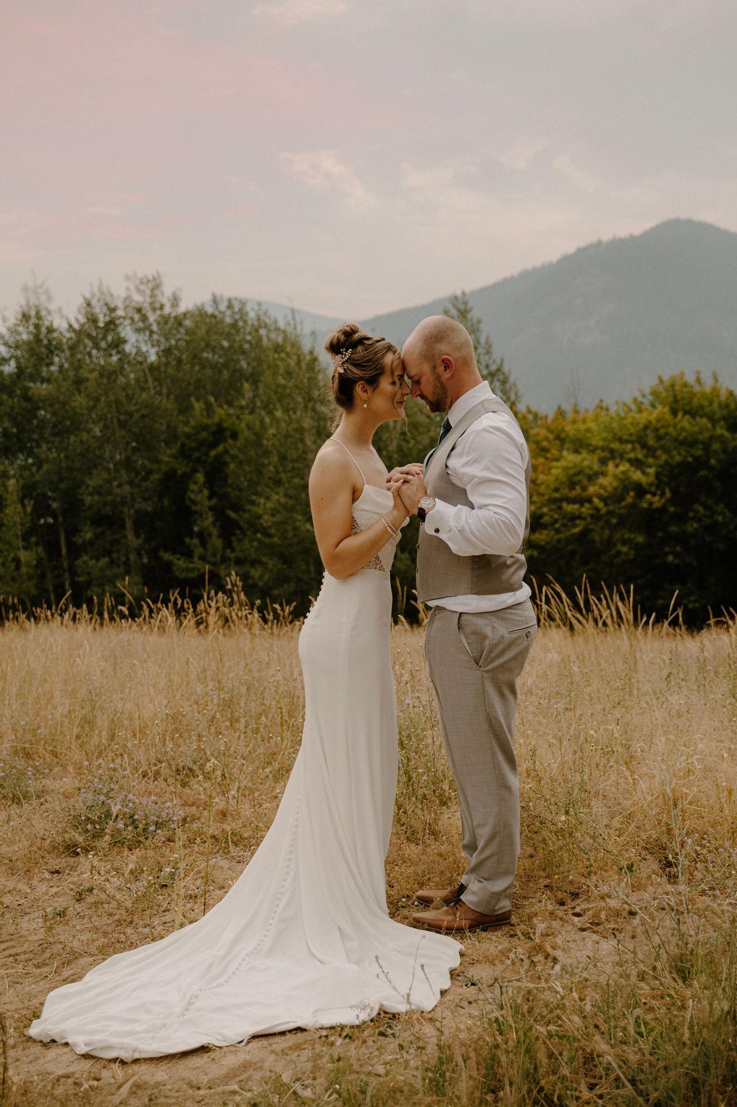 Bride and groom holding hands in a golden field with mountain and forest backdrop at sunset.