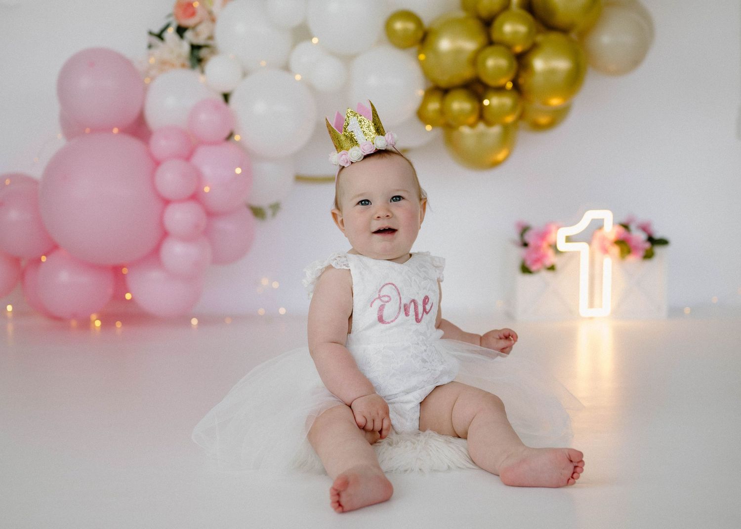 Baby girl wearing gold crown sits on floor in white One romper at first birthday with pink balloon garland.