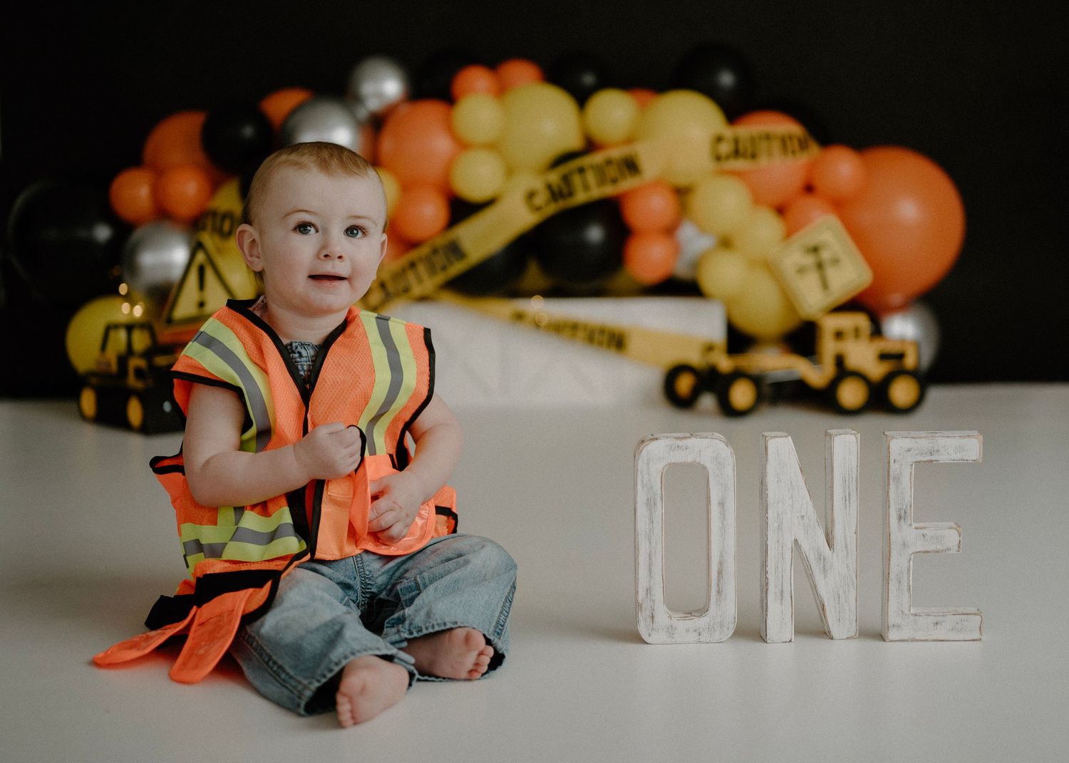Baby boy in construction vest sits beside ONE letters with balloon garland backdrop.
