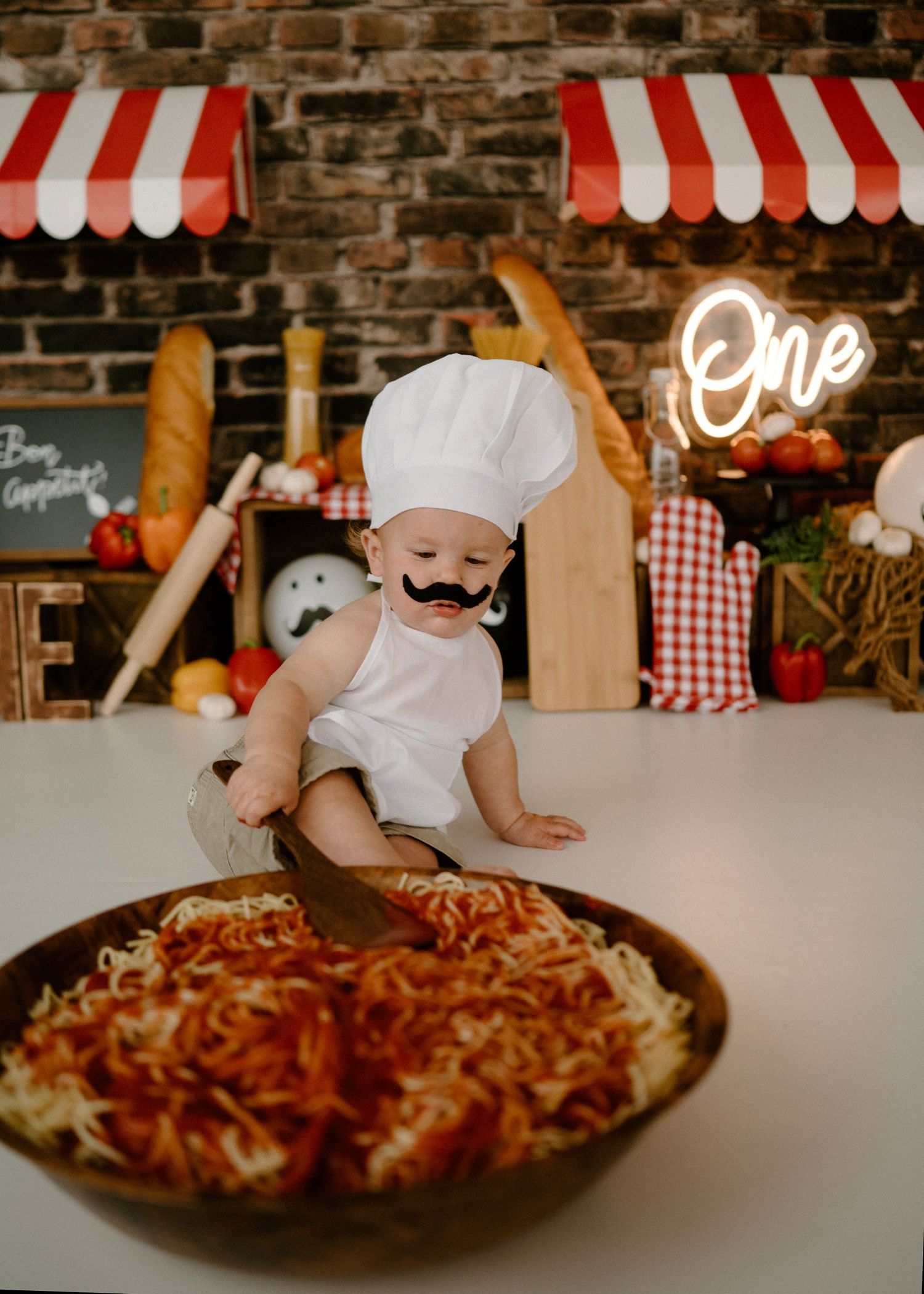 Baby with fake mustache and chef hat reaches into spaghetti bowl at Italian-themed first birthday session.