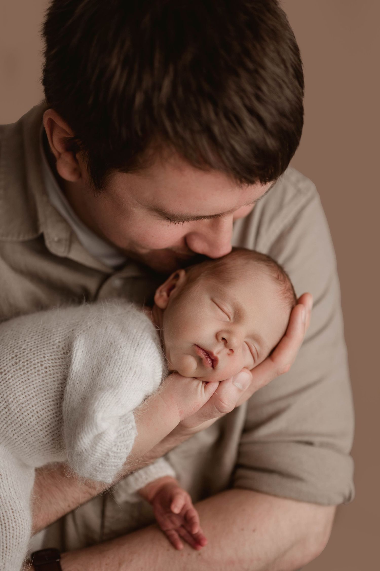 Father gently kissing sleeping newborn baby wrapped in white, cradled in his hands.