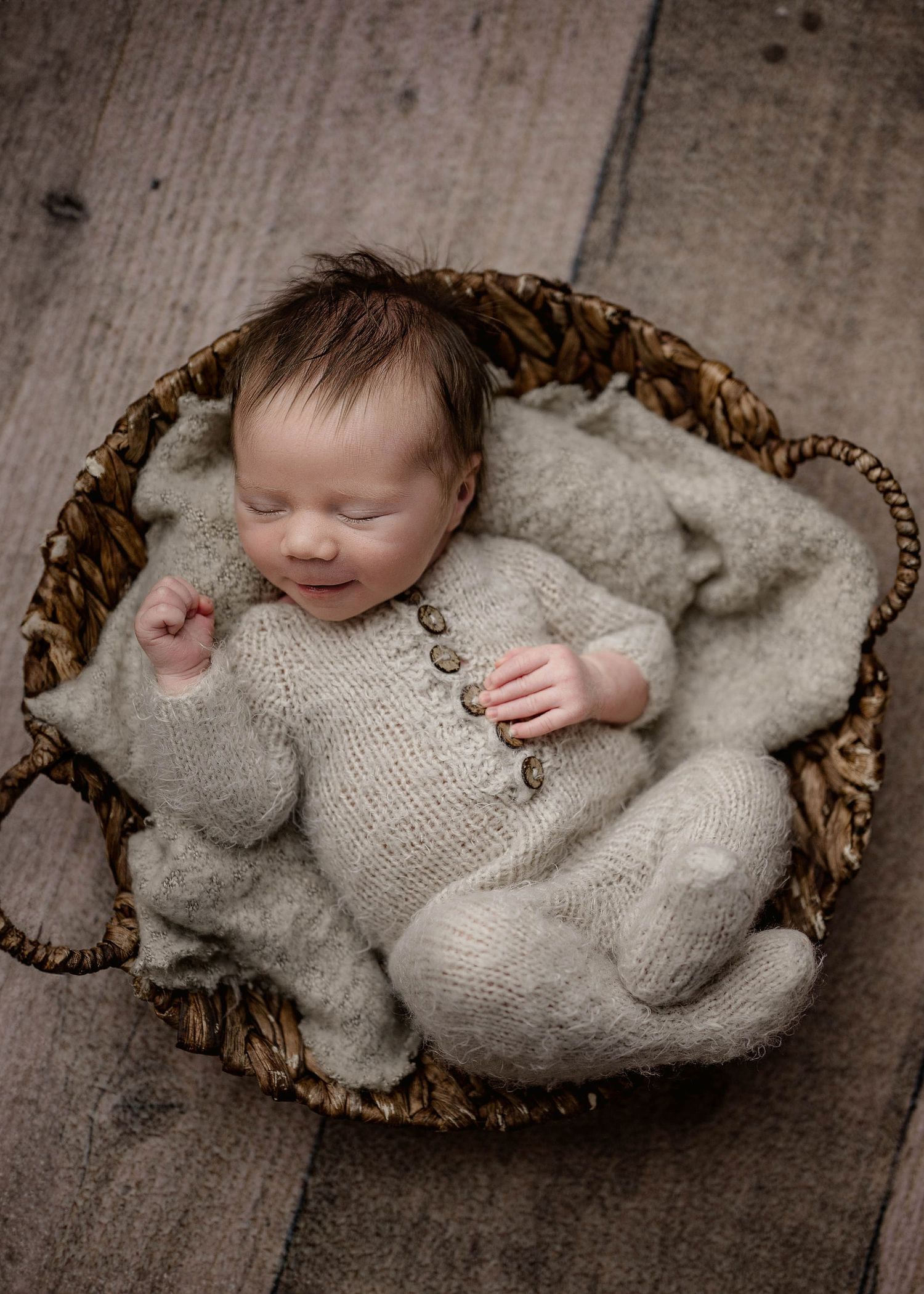 Sleeping newborn baby in a knit gray romper nestled in a woven basket on a rustic wood floor in Leduc.