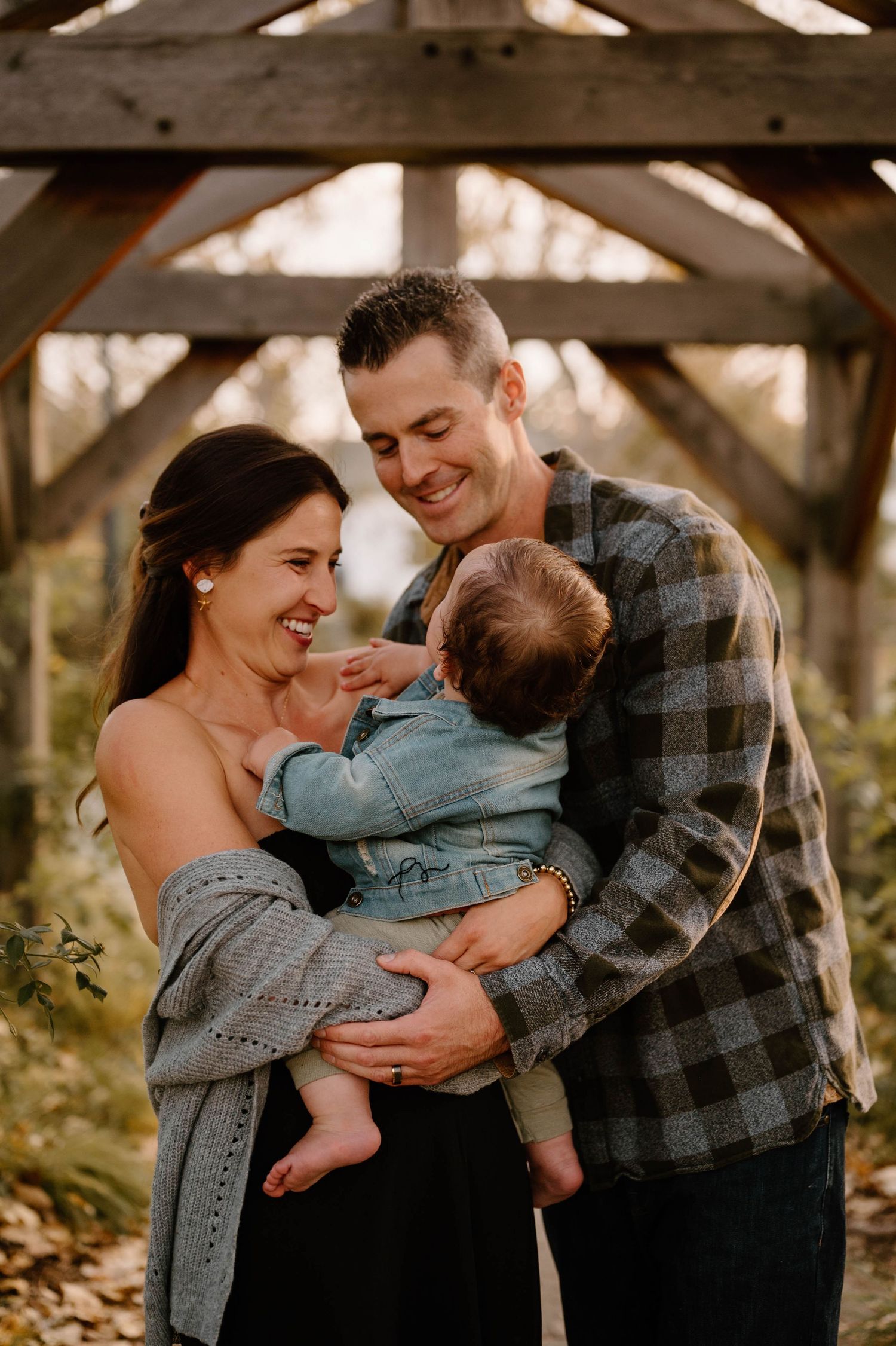 Smiling parents bonding with their baby boy under rustic wooden pergola in fall garden setting.