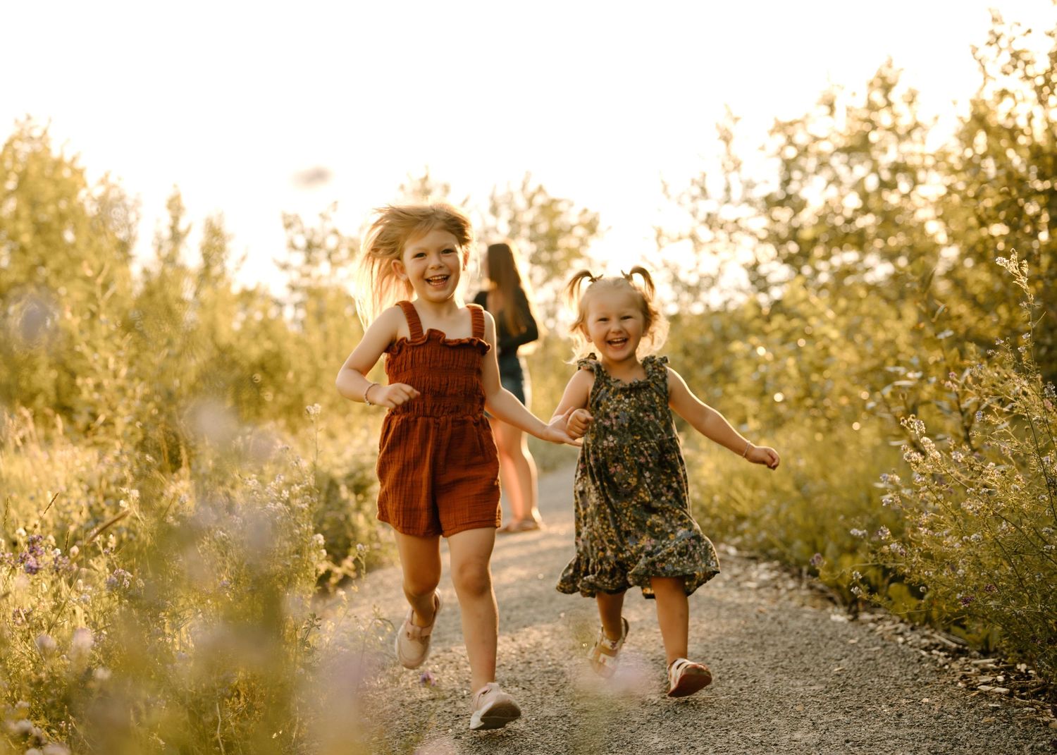 Two young sisters in colorful dresses holding hands and running on a sunny path.