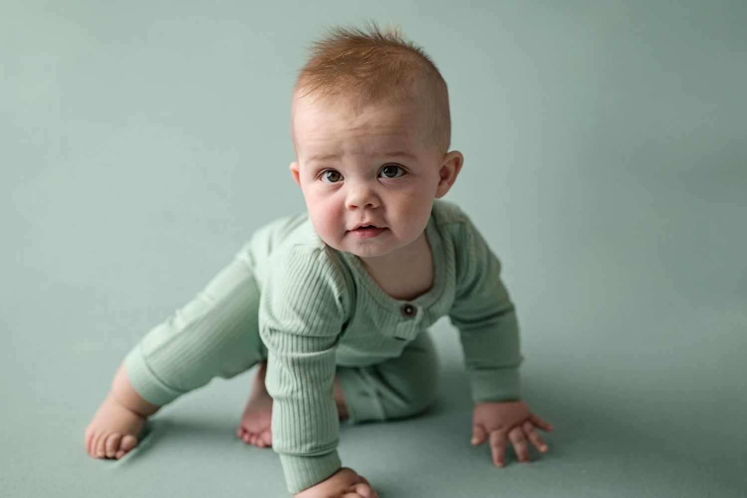 Adorable red-haired infant in sage green ribbed outfit crawling and looking at camera in studio portrait.