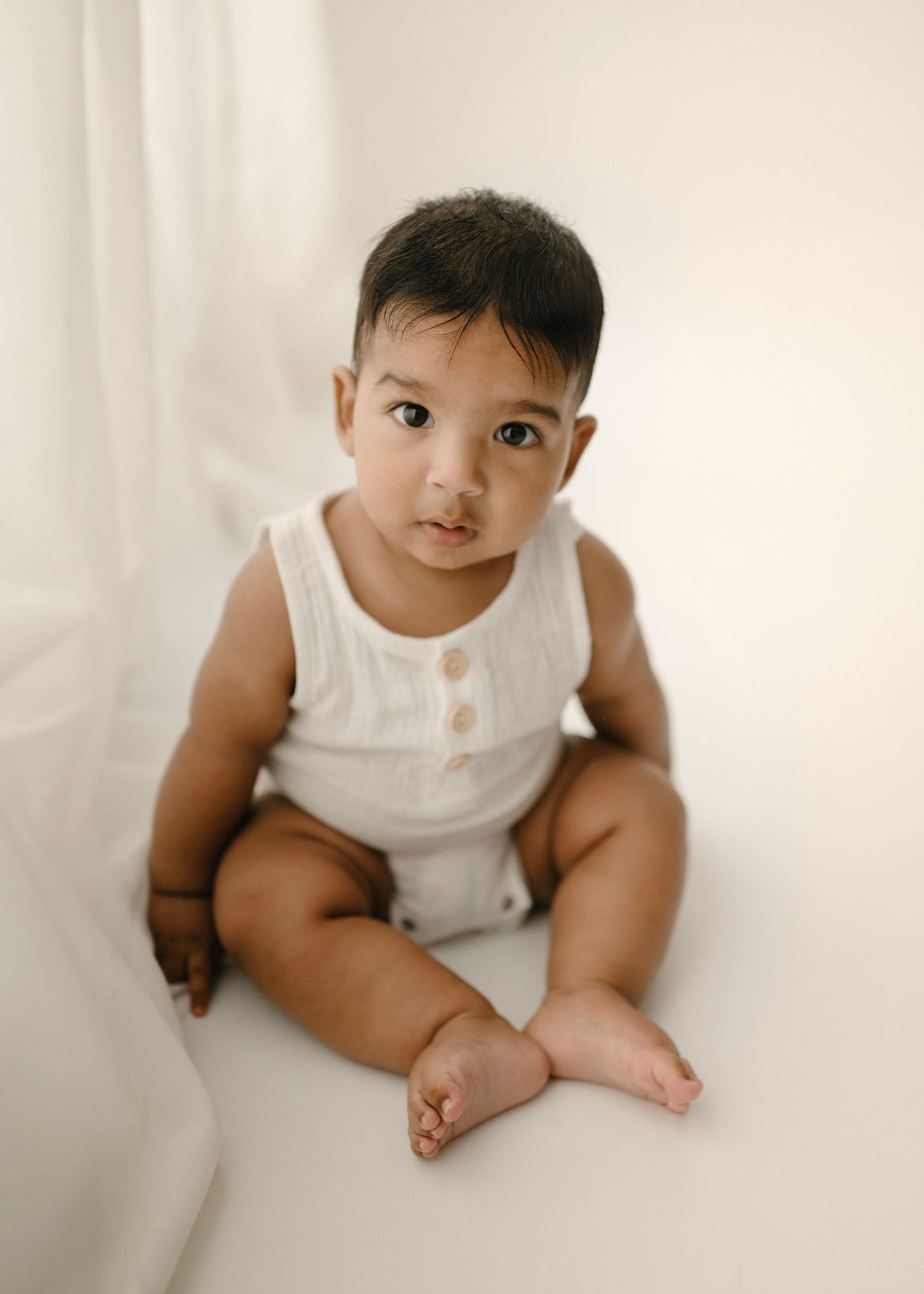 Curious baby in white linen romper sitting on white background.