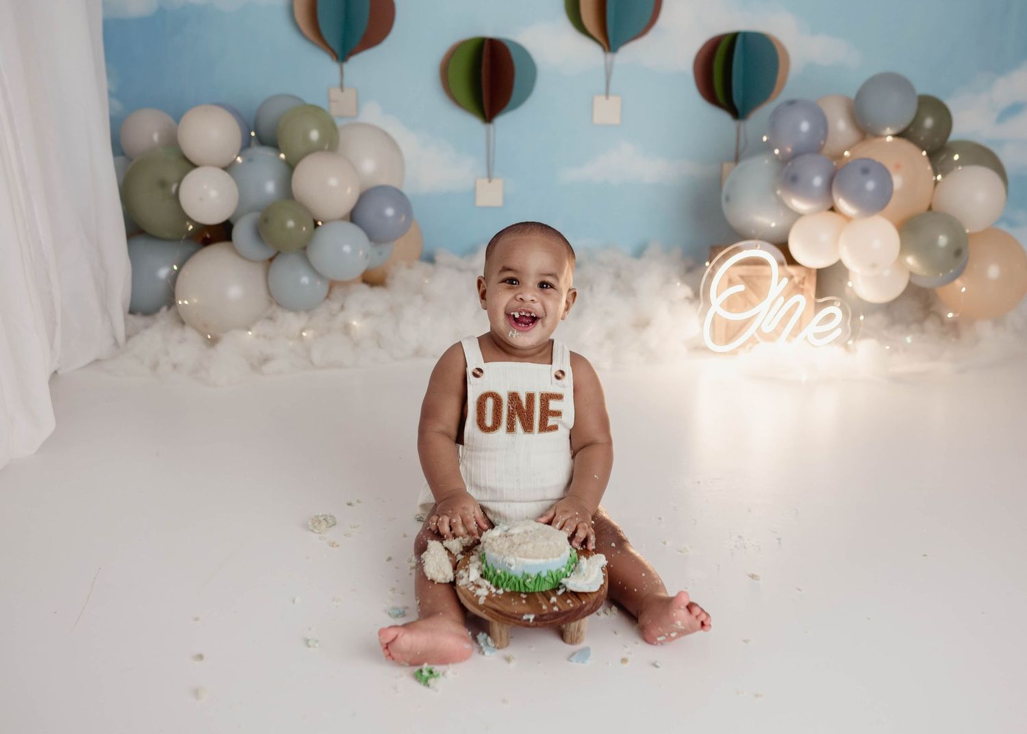 Happy baby boy celebrating first birthday with cake smash, wearing ONE romper with balloon backdrop.