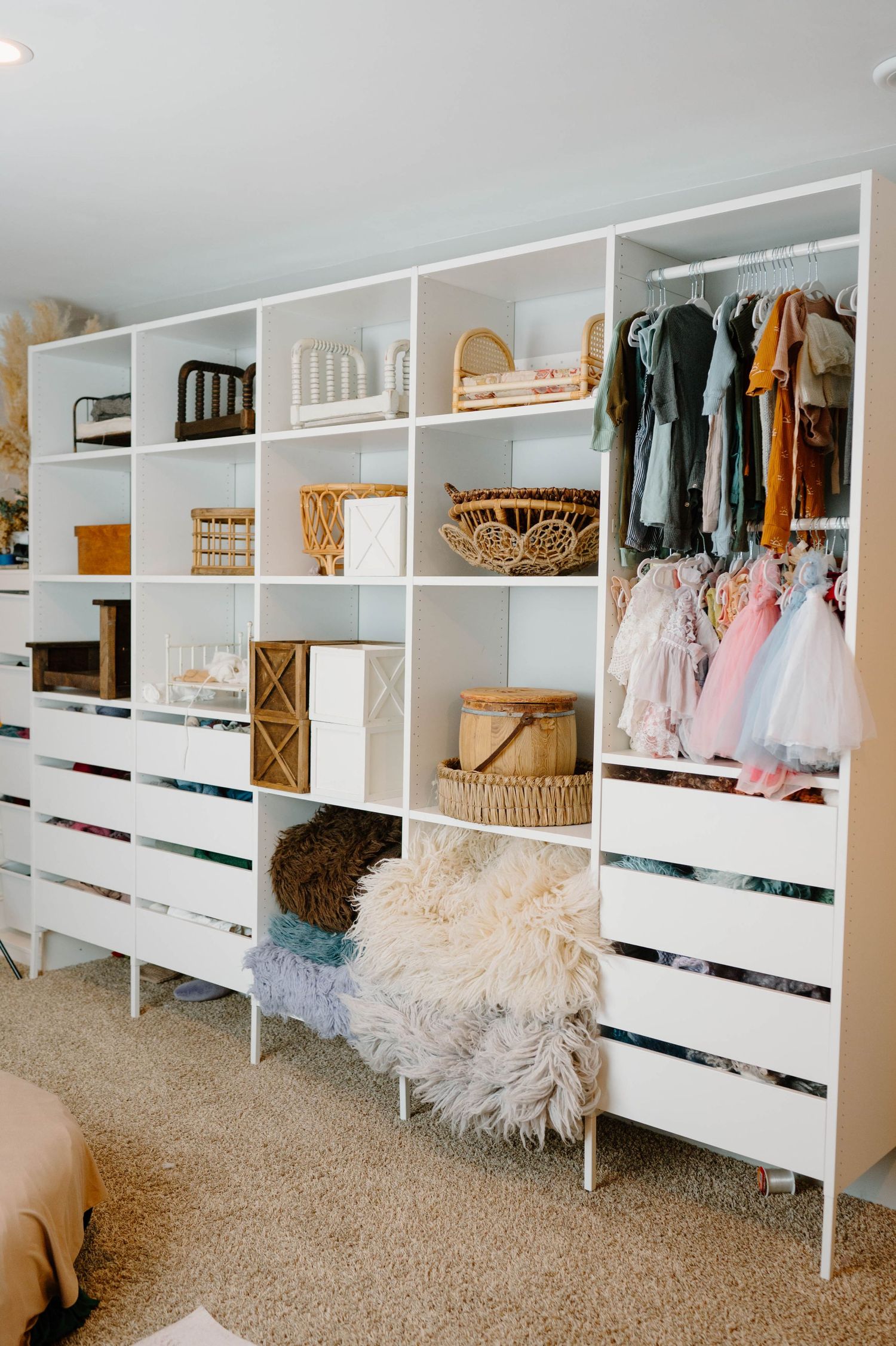 Large white IKEA-style shelving unit with wicker baskets, props, hanging children's dresses, and drawers.