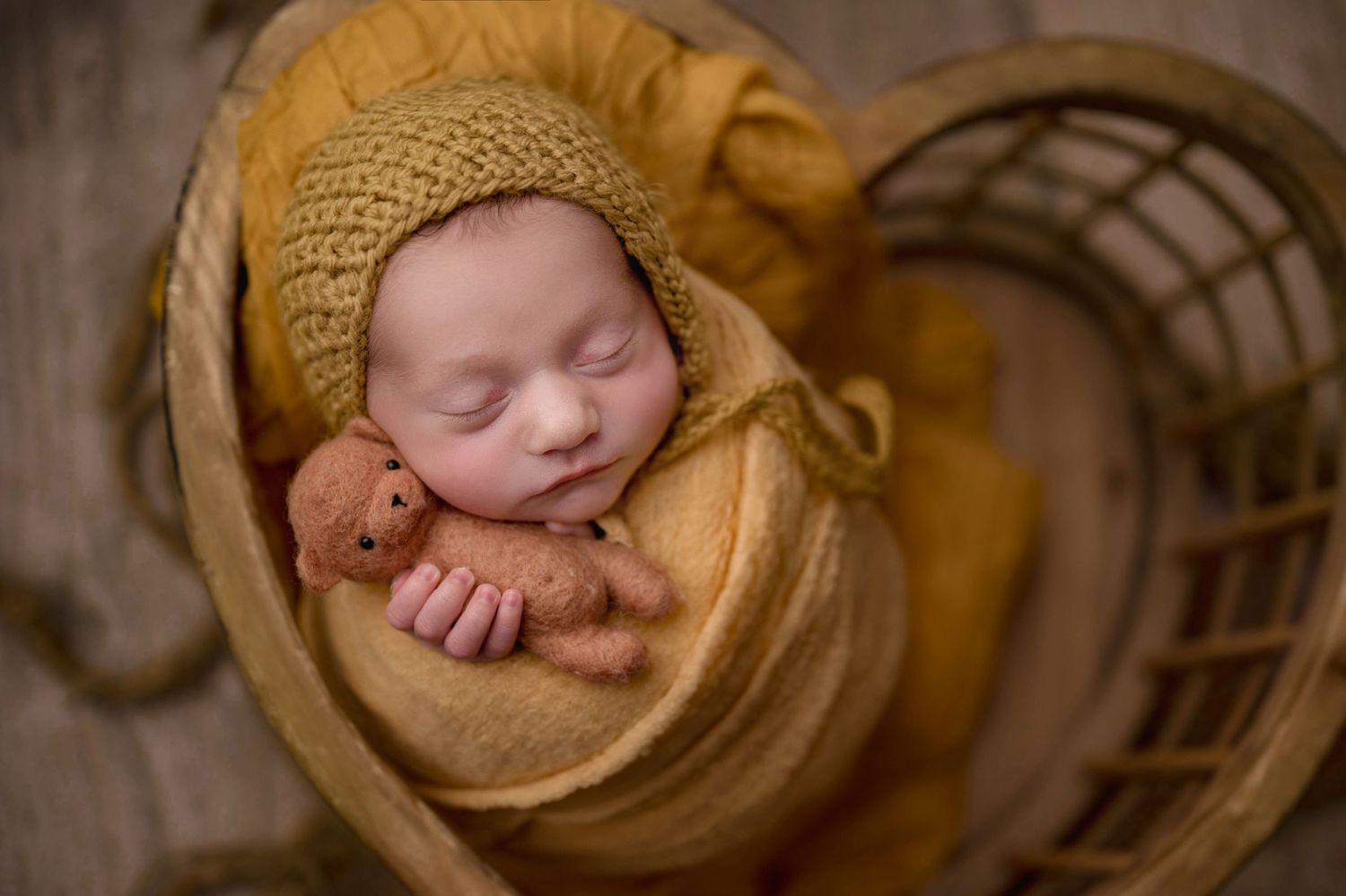 Newborn baby sleeping in golden yellow wrap and knit bonnet, cradled in wicker basket holding tiny teddy bear.