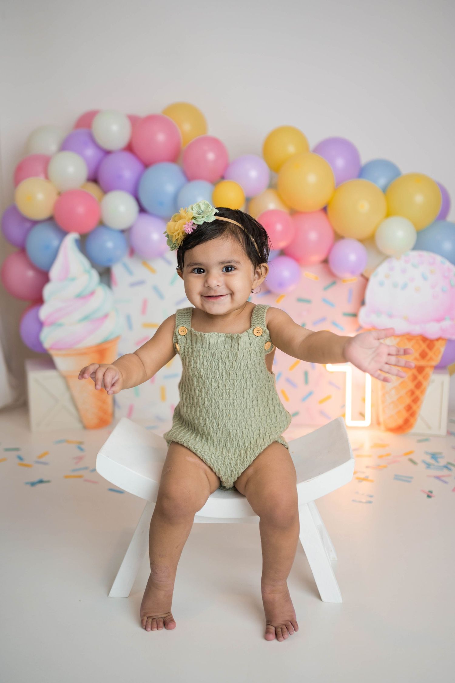 Happy baby girl in green romper sits smiling at ice cream themed first birthday photo session.