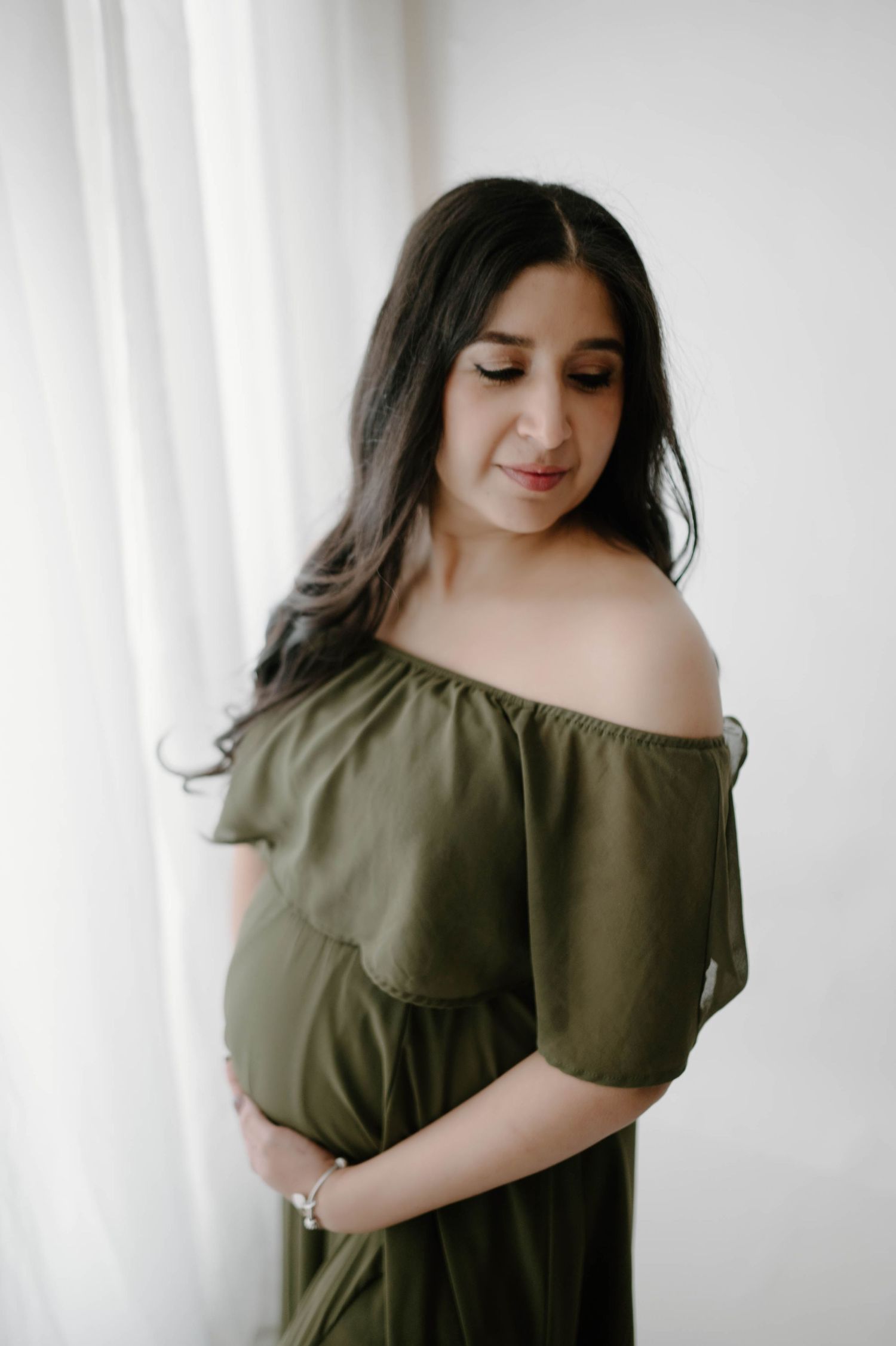 Pregnant woman in olive off-shoulder dress gazing down softly near white curtains.