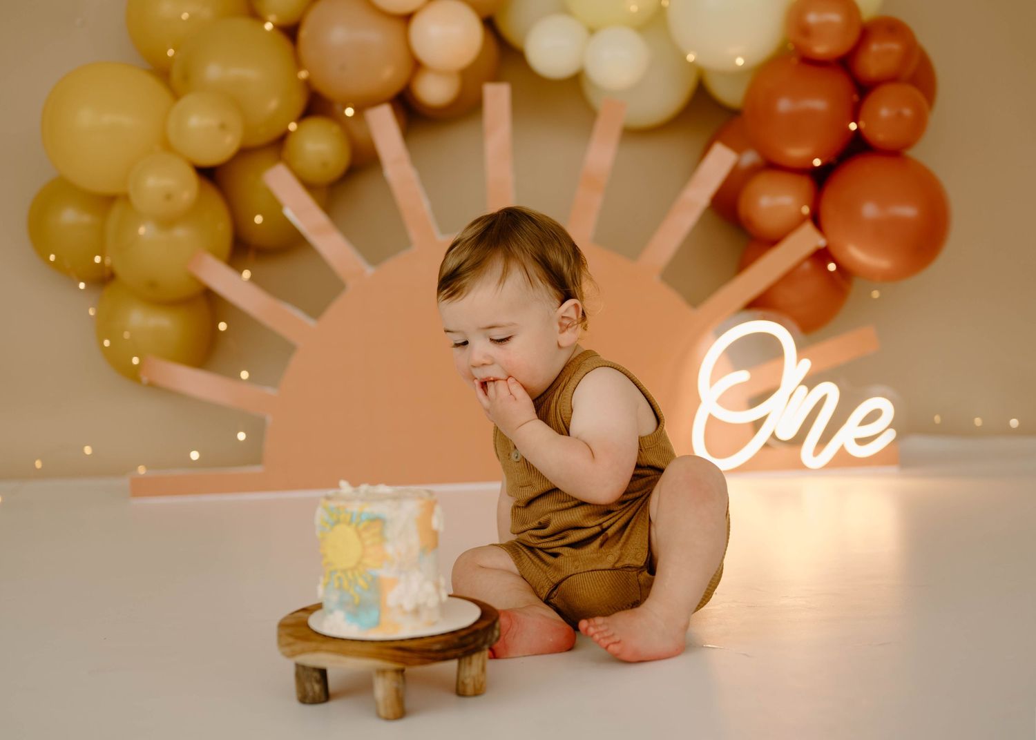 One year old baby boy smashing first birthday cake with boho rainbow balloon arch backdrop.