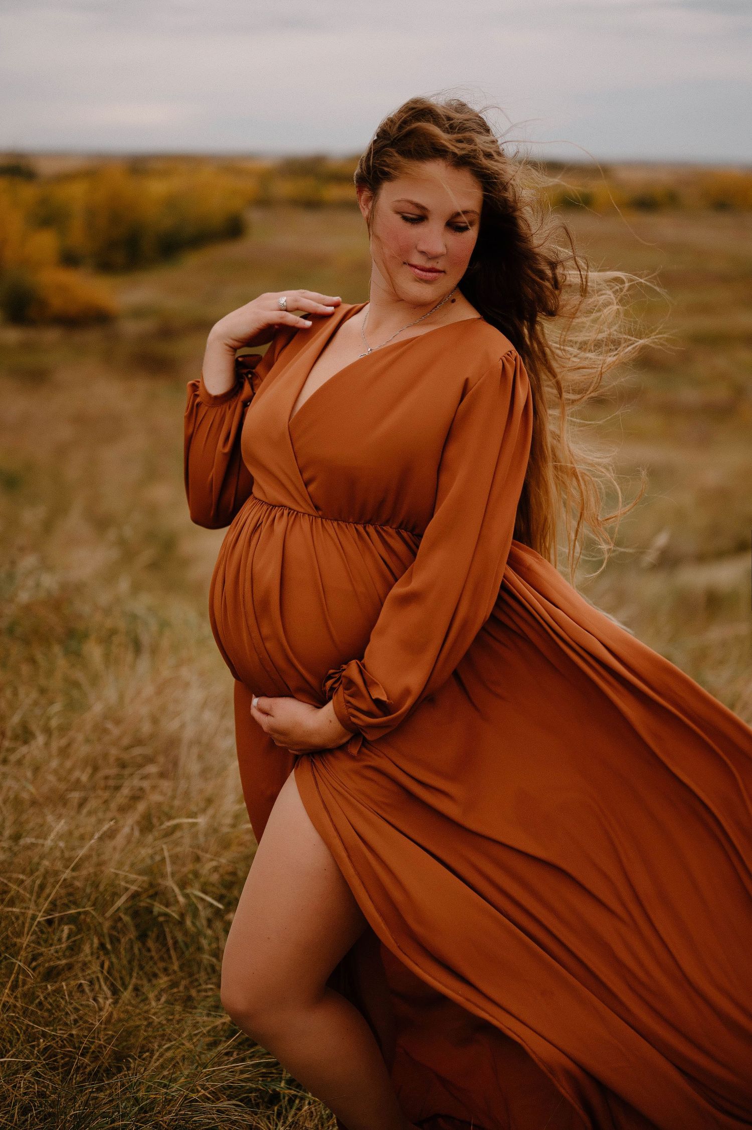 A pregnant woman in a flowing rust wrap dress cradles her bump while posing in an grassy field near the LRC.
