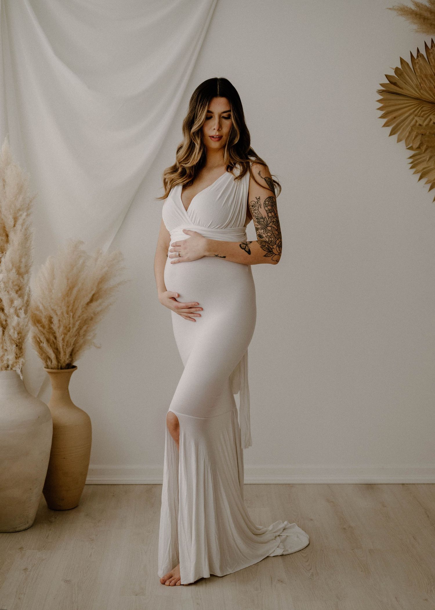 Pregnant woman in white fitted gown with tattoos posing in a boho studio.