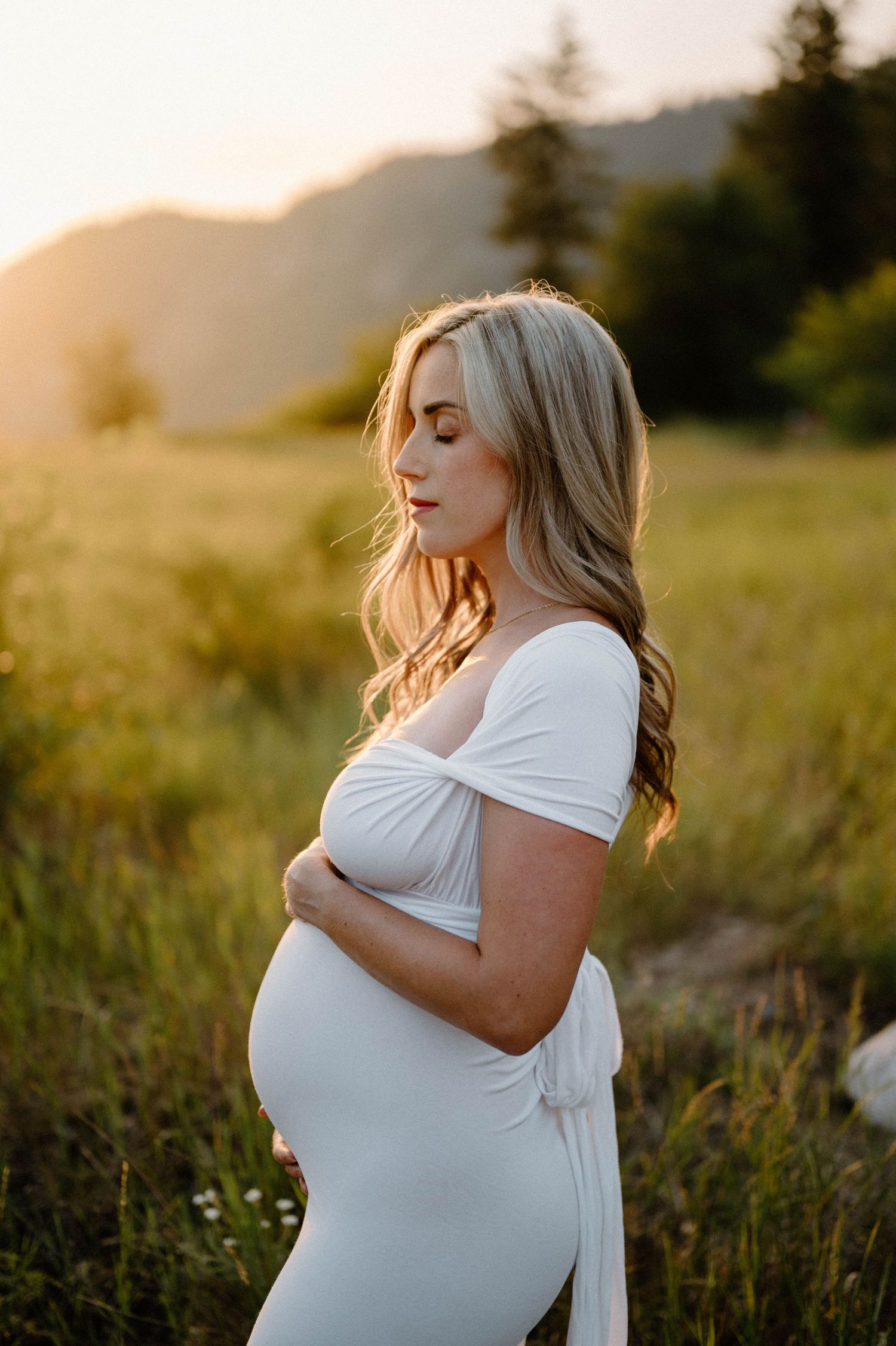 Pregnant woman in white off-shoulder dress holds her belly in a sunlit mountain meadow at sunset.