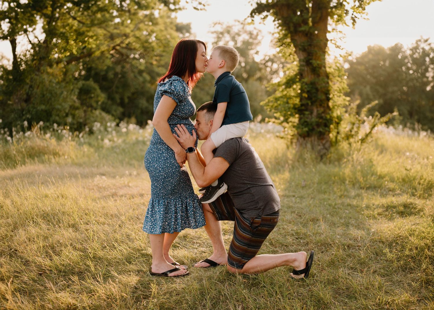 Pregnant woman and partner share a kiss while lifting their young child in a sunny green outdoor setting.
