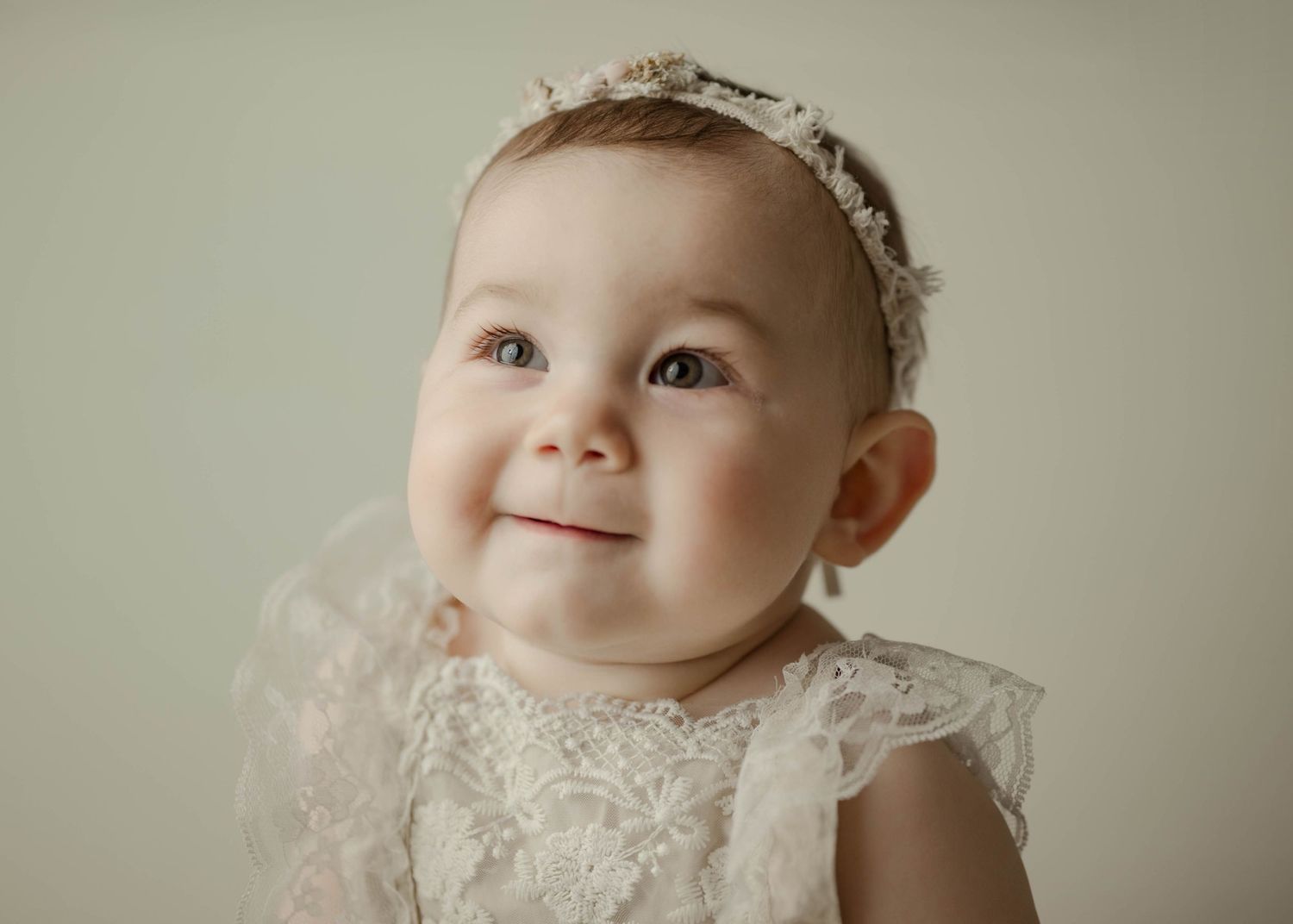 Adorable baby girl in white lace dress and floral headband smiling softly against neutral background.