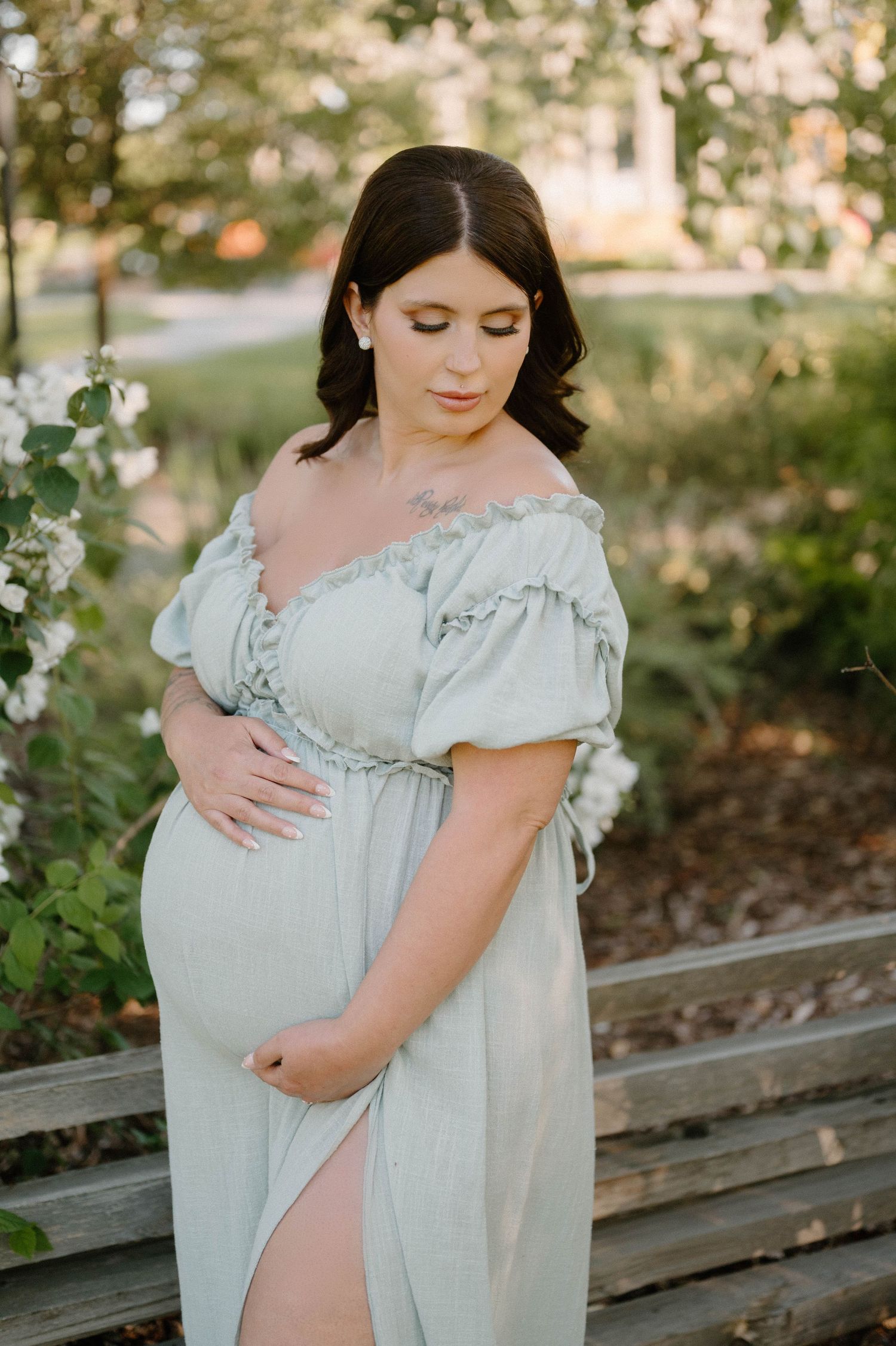 Pregnant woman in sage green off-shoulder dress cradling baby bump outdoors near flowering bushes.
