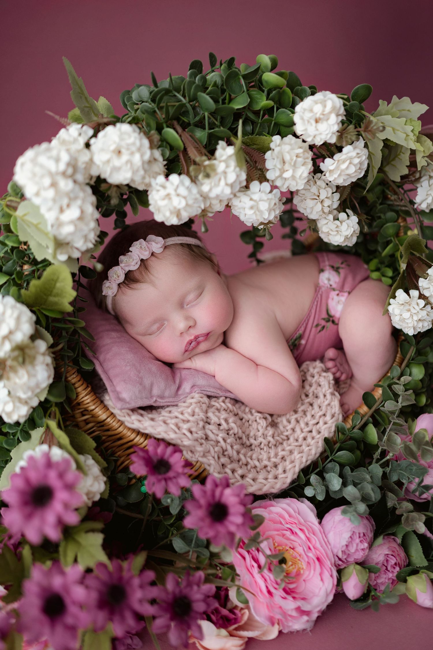 Sleeping newborn baby in floral wreath basket with white and pink flowers on pink background.
