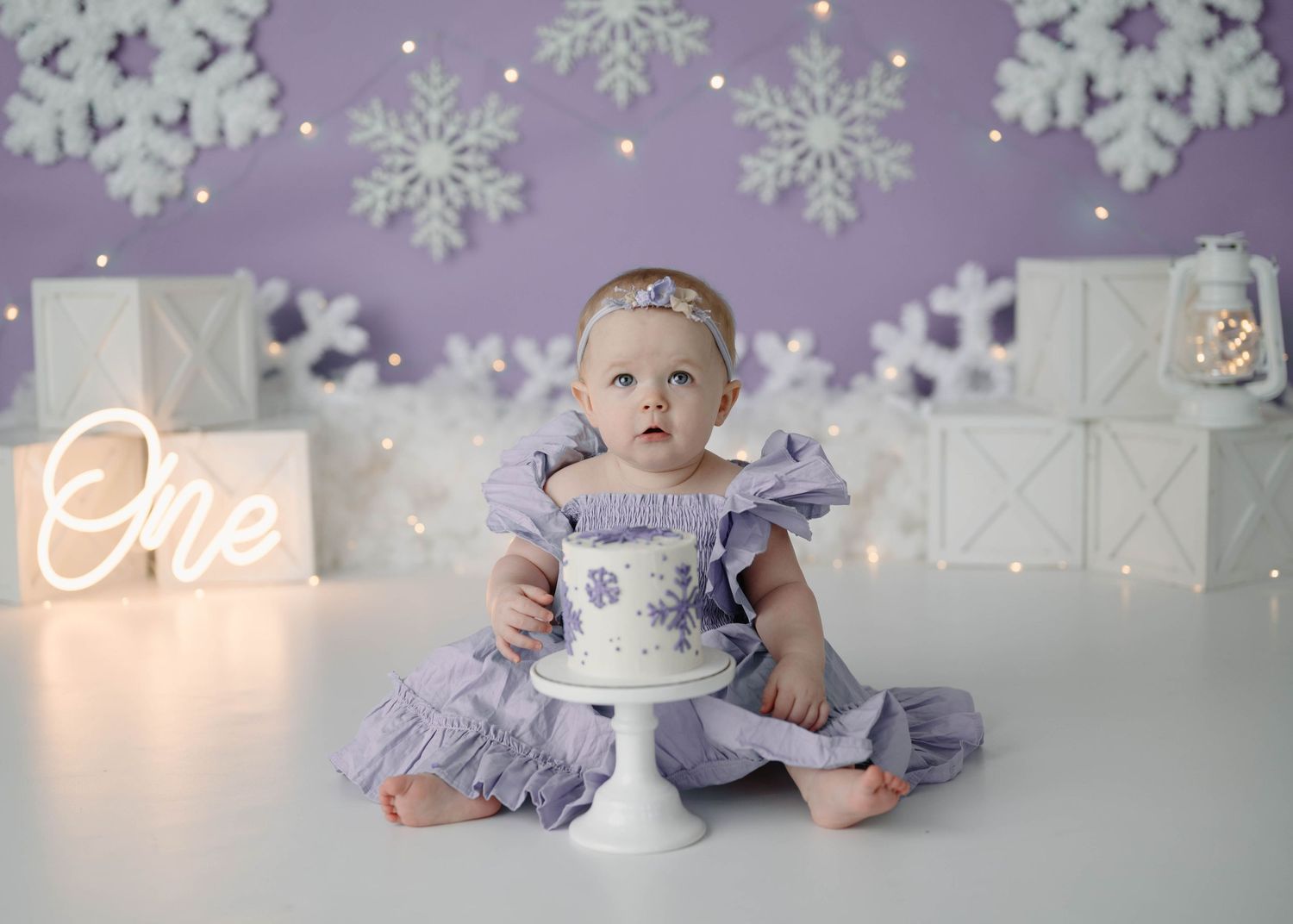 Baby girl in lavender ruffle dress sits before decorated first birthday cake with snowflake theme backdrop.