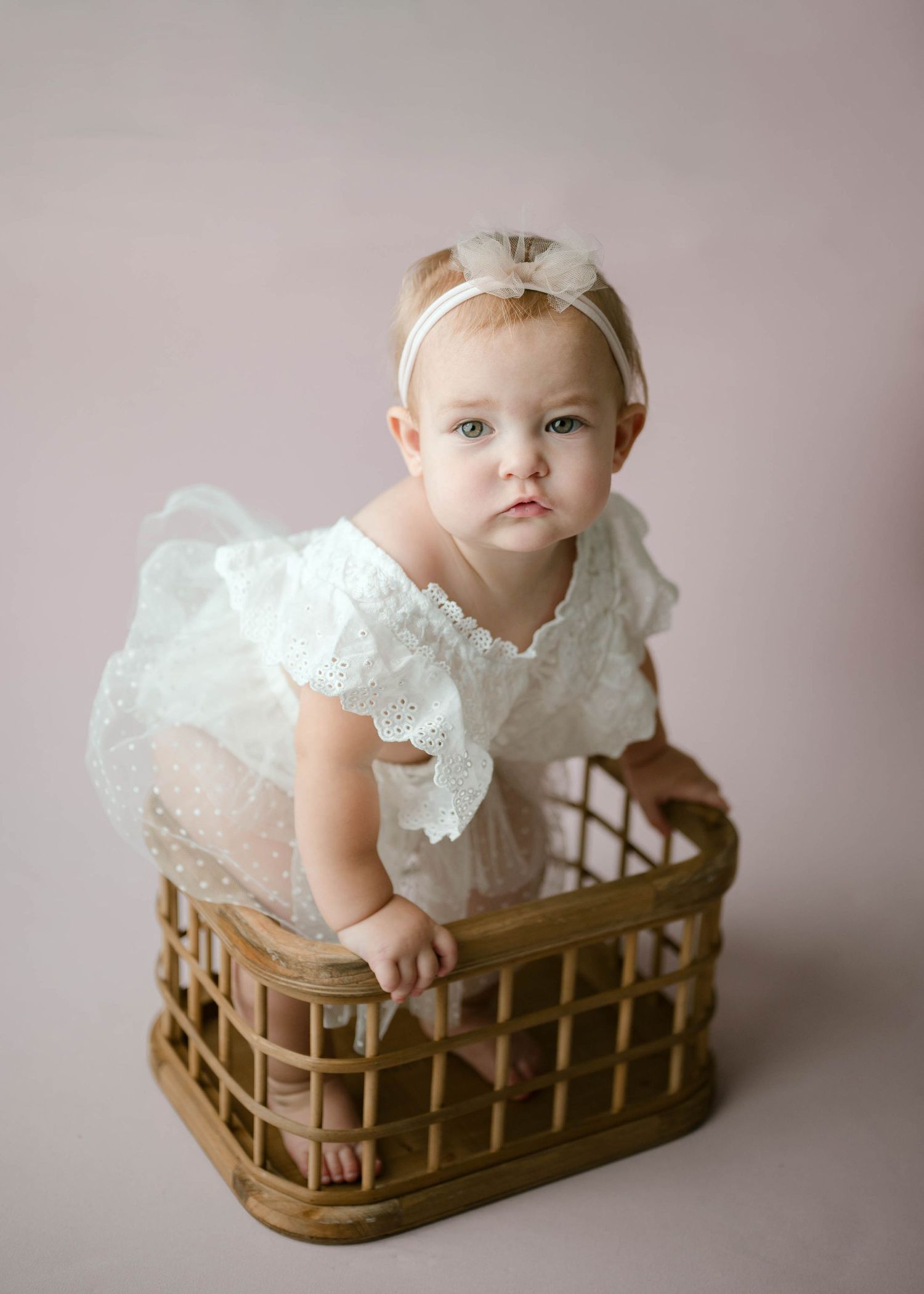 Baby girl in white lace romper with bow headband stands in wicker basket against pink studio backdrop.