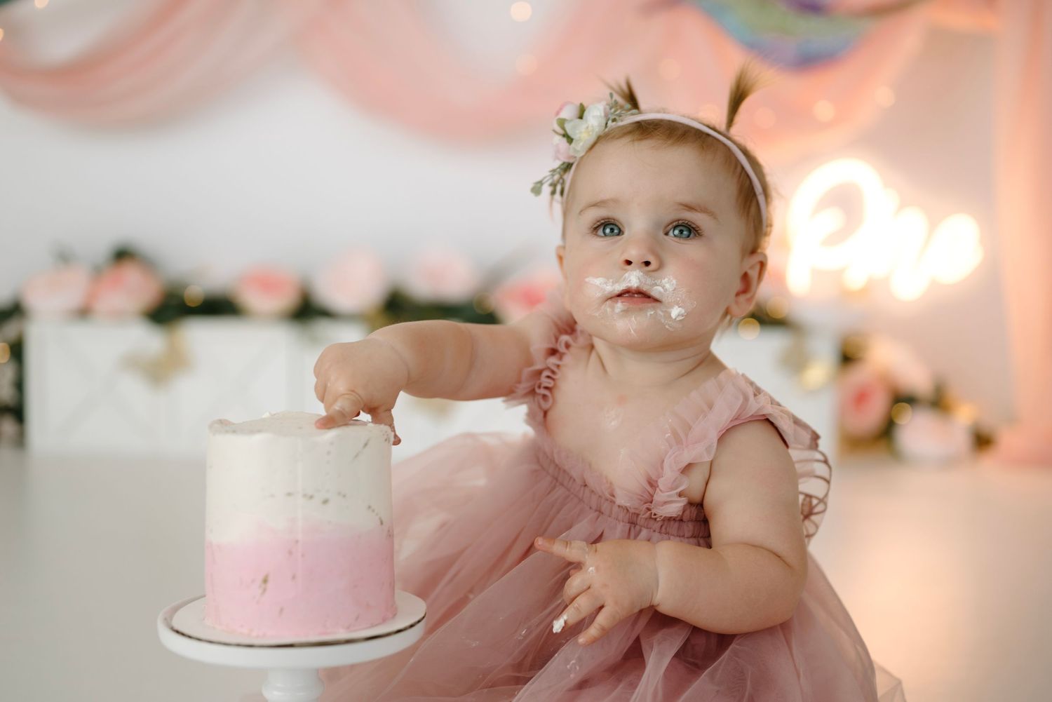 Baby girl in pink ruffle dress reaches for a pink ombre cake at a floral hummingbird first birthday session.