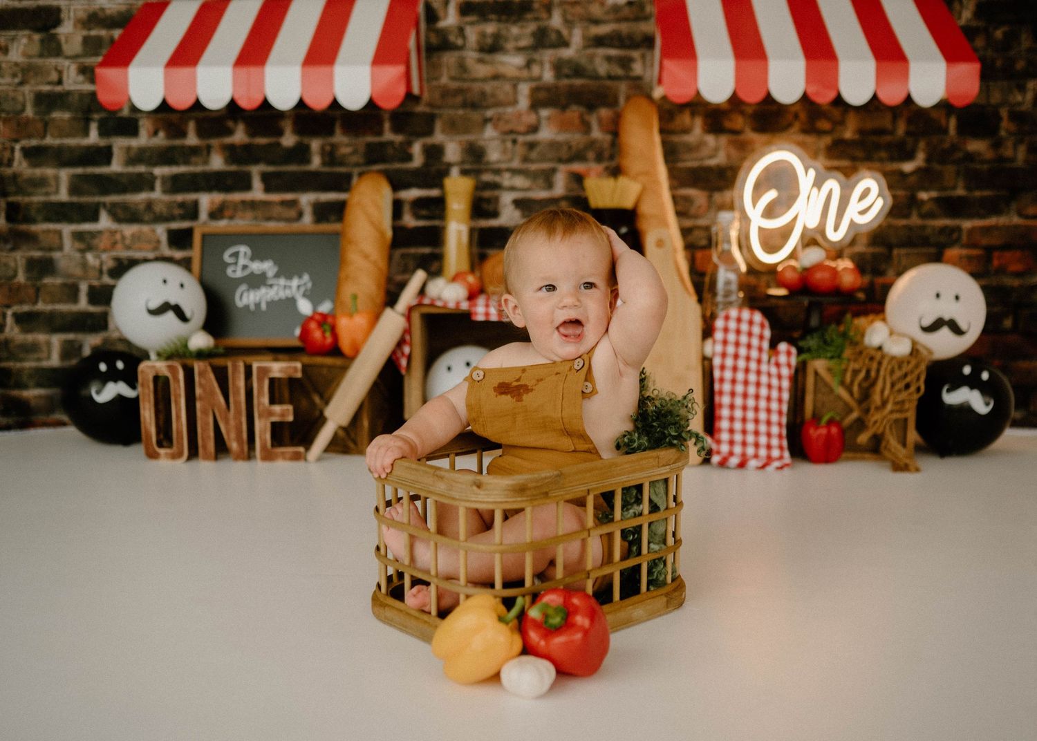 Baby in brown overalls sits in wooden crate with fresh vegetables at Italian-themed birthday session.