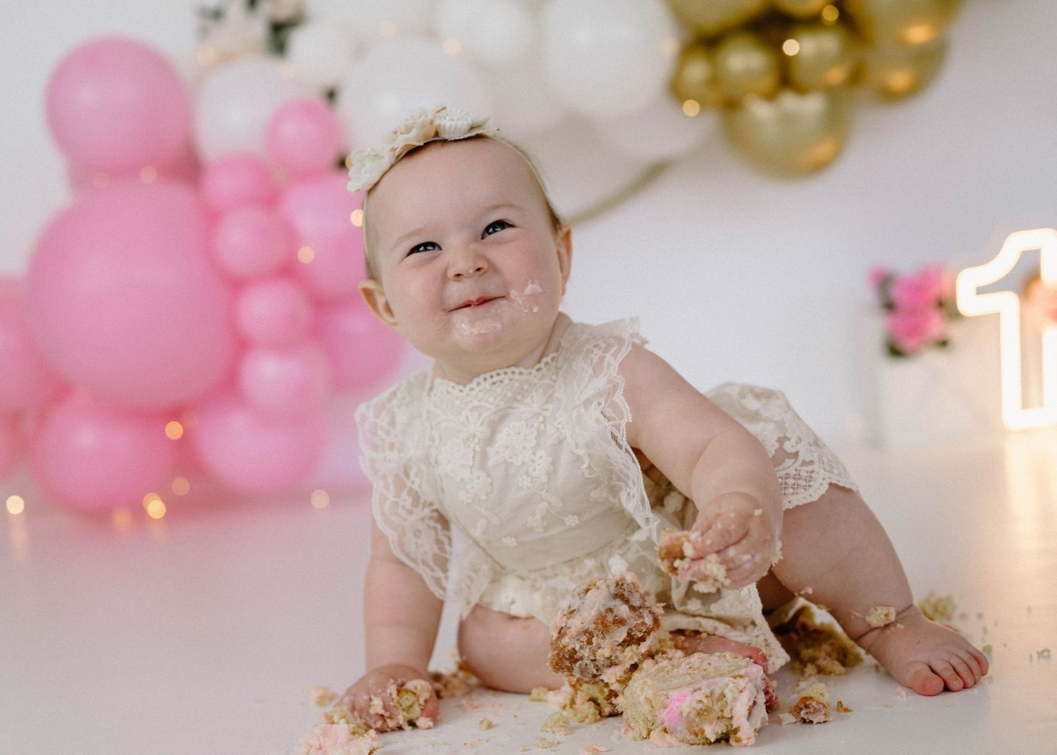 Smiling baby girl in lace outfit celebrates first birthday with smashed cake and pink and gold balloons.