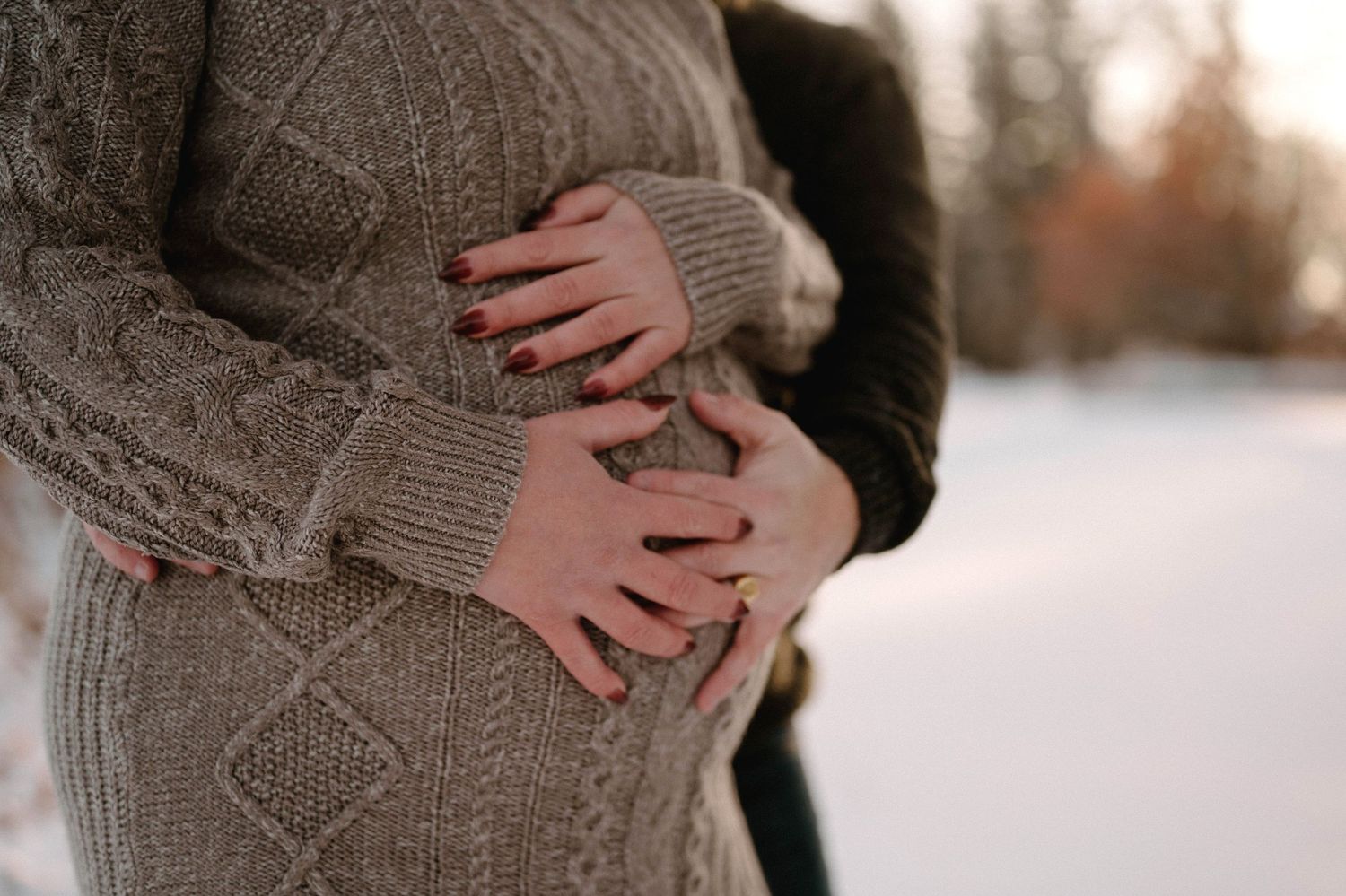 Close-up of couple holding pregnant belly together outdoors in snowy winter setting.